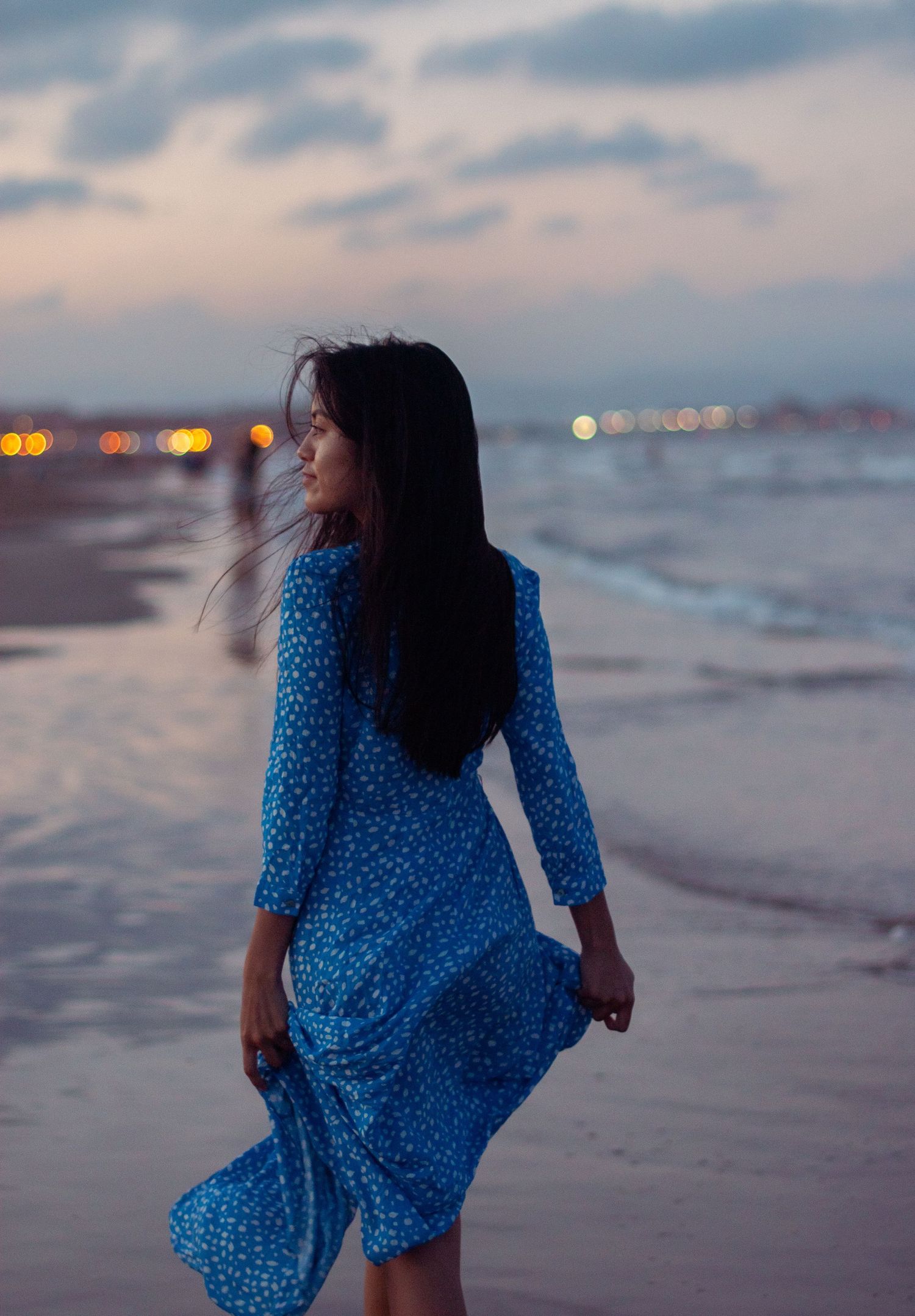 A person in flowing blue polka dot dress walks along shoreline at dusk.