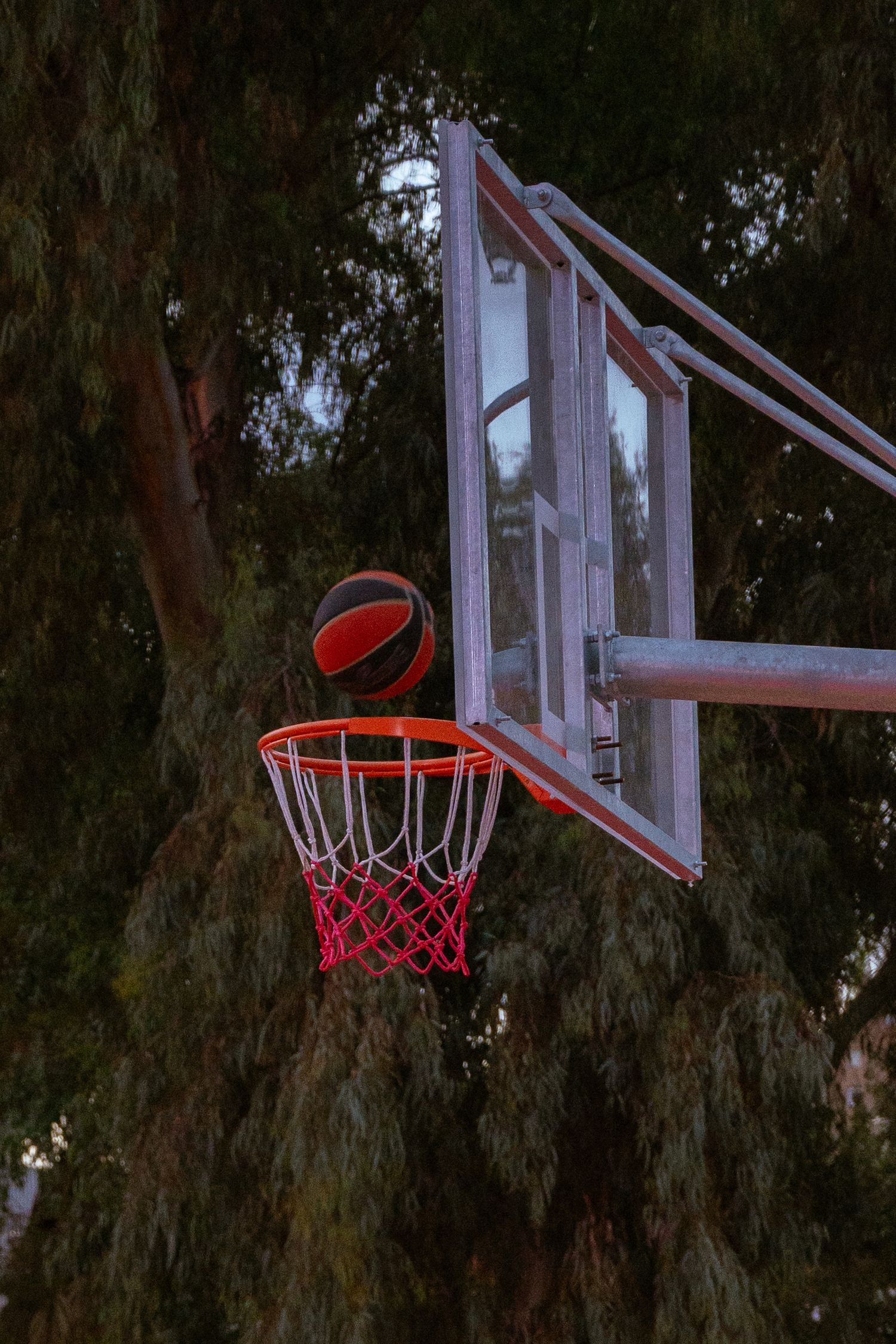 A basketball hoop with red netting stands against a dark background of trees at dusk.