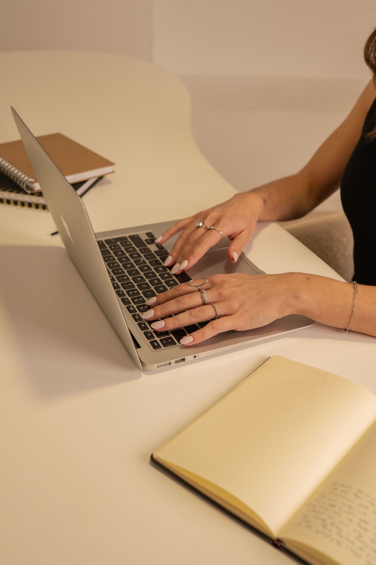 Hands typing on laptop keyboard with notebook nearby on white surface.
