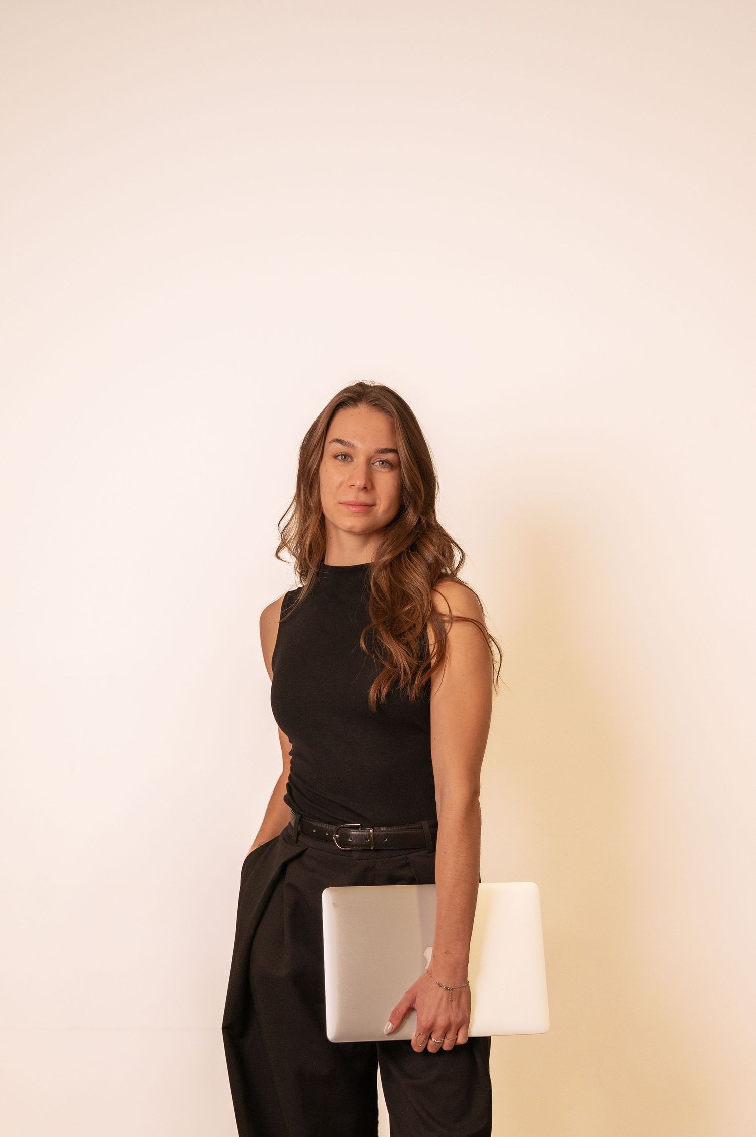 A professional woman in black sleeveless top and trousers holding a laptop against a neutral background.