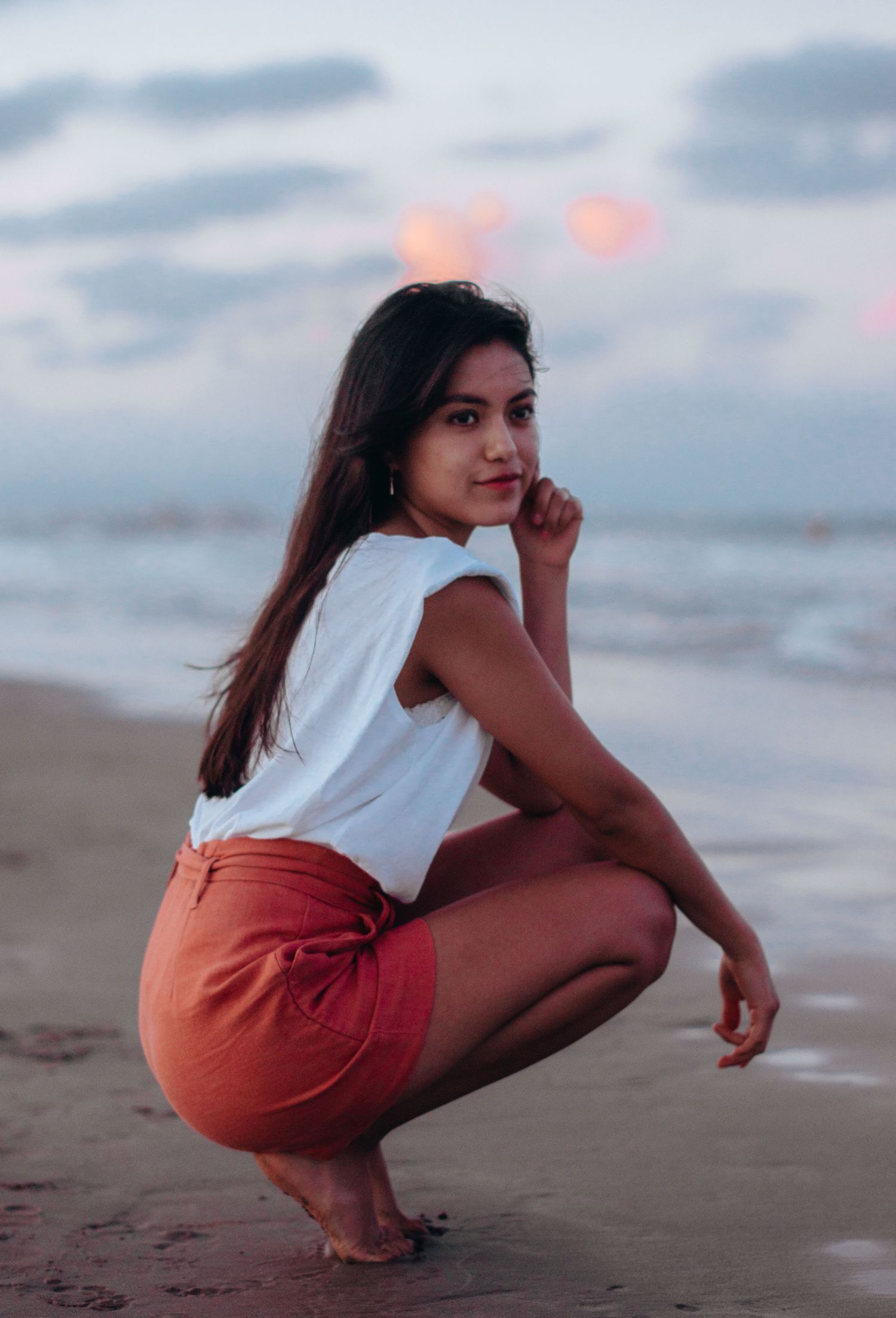 A person in coral shorts and white top crouches on the beach at twilight.