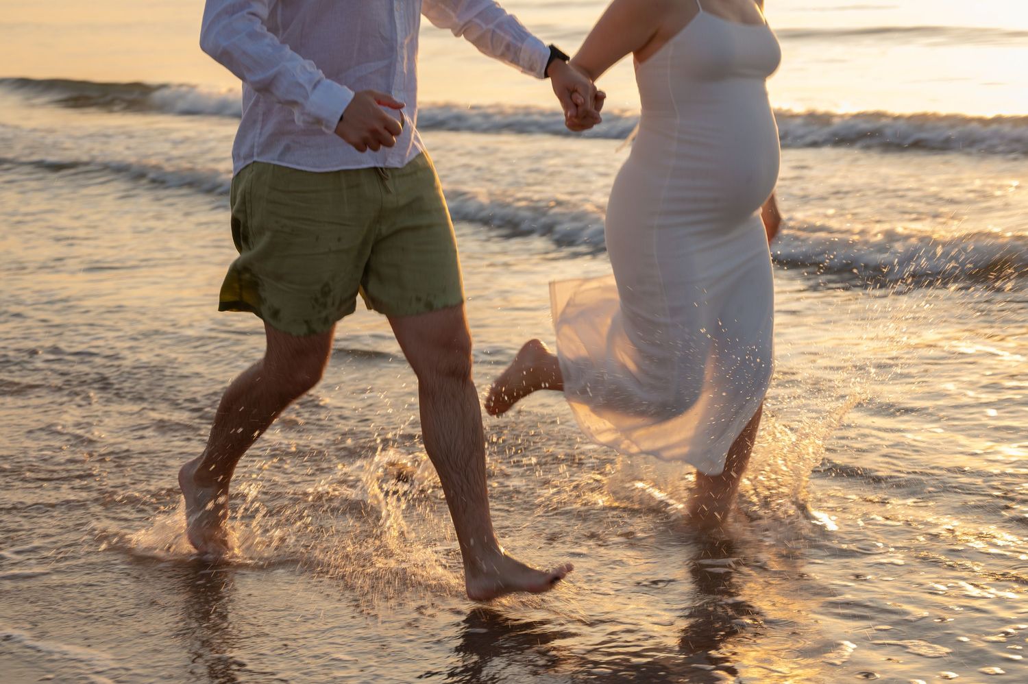 Pregnant couple running hand-in-hand through ocean waves on a beach at golden sunset.
