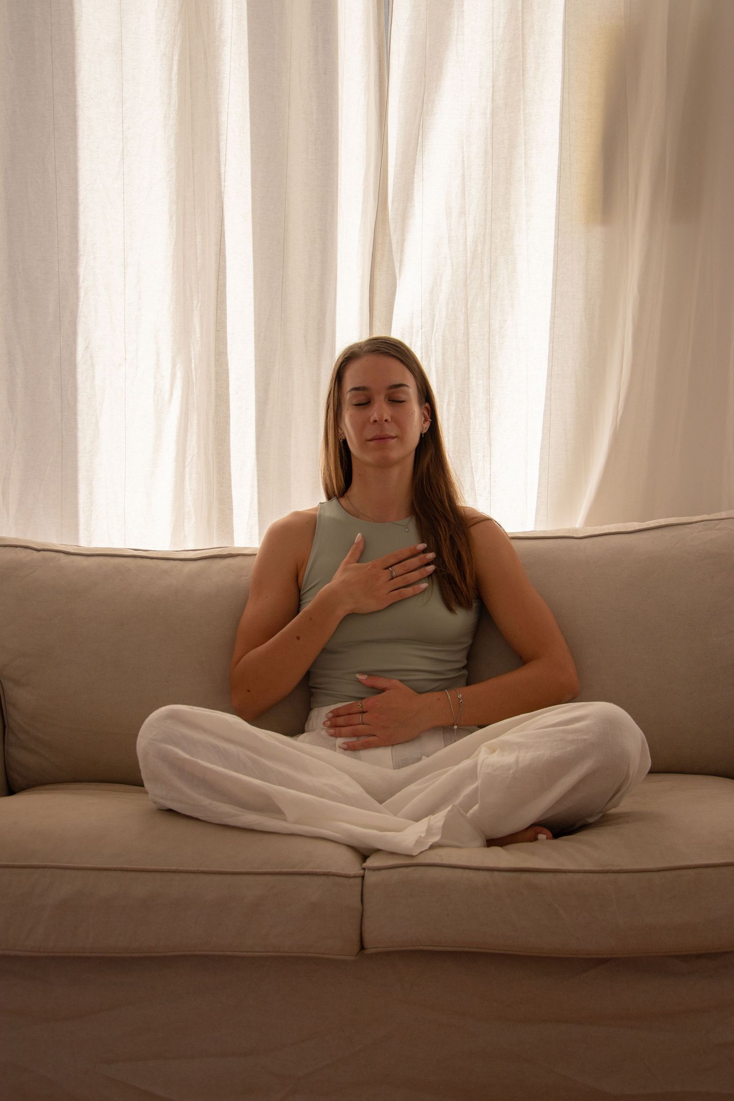 Person in meditation pose sitting cross-legged on grey couch in sunlit room.