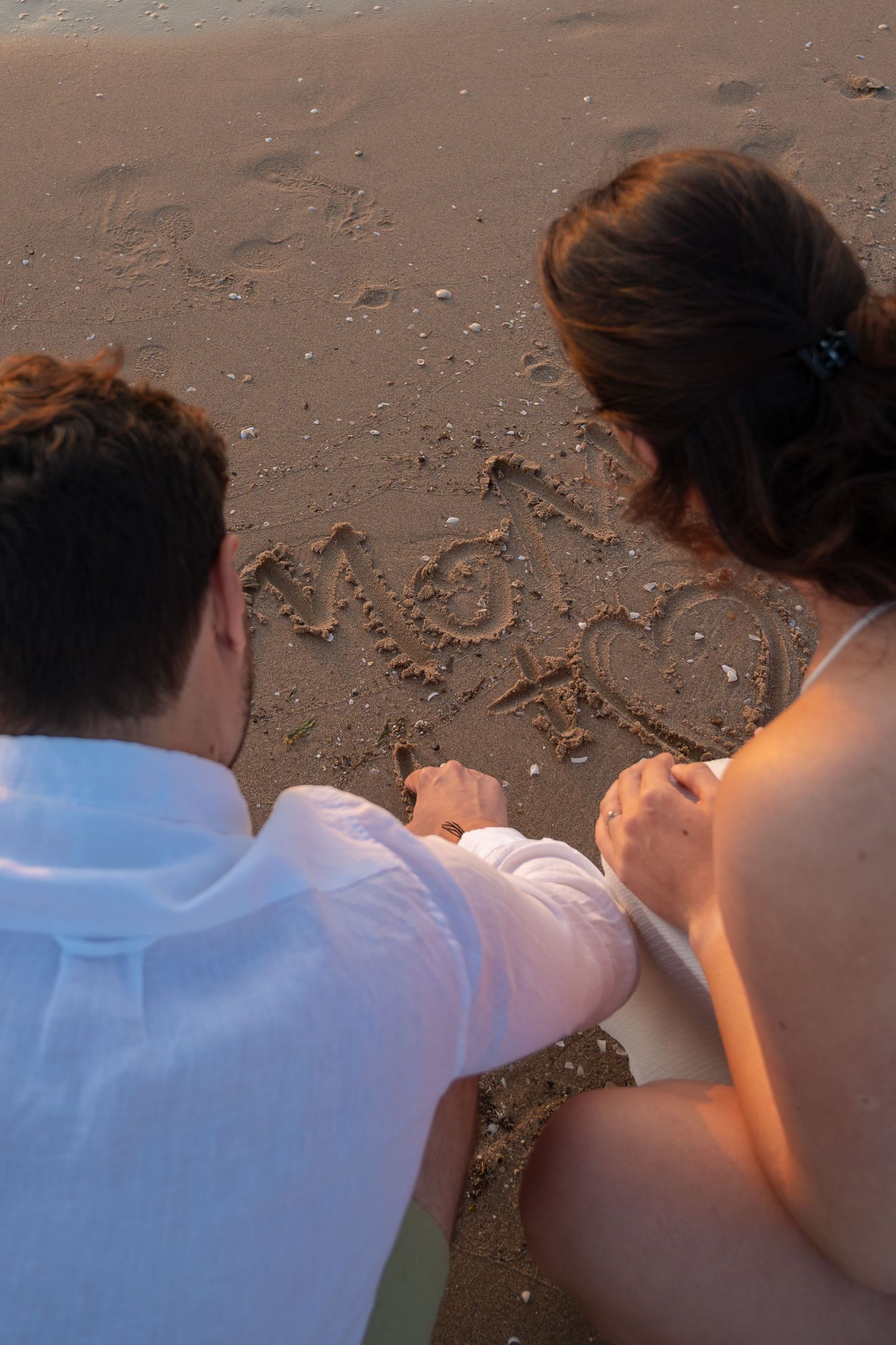 A couple draws a heart in the sand on a beach during golden hour.