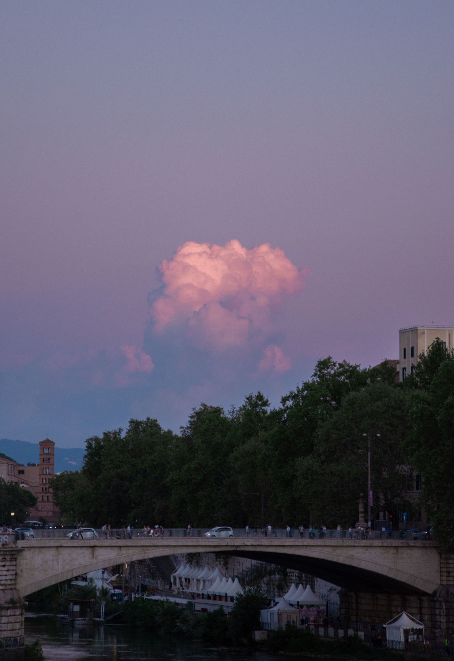 A stunning pink sunset sky with a glowing cumulus cloud above a stone bridge over a calm river in a European city.