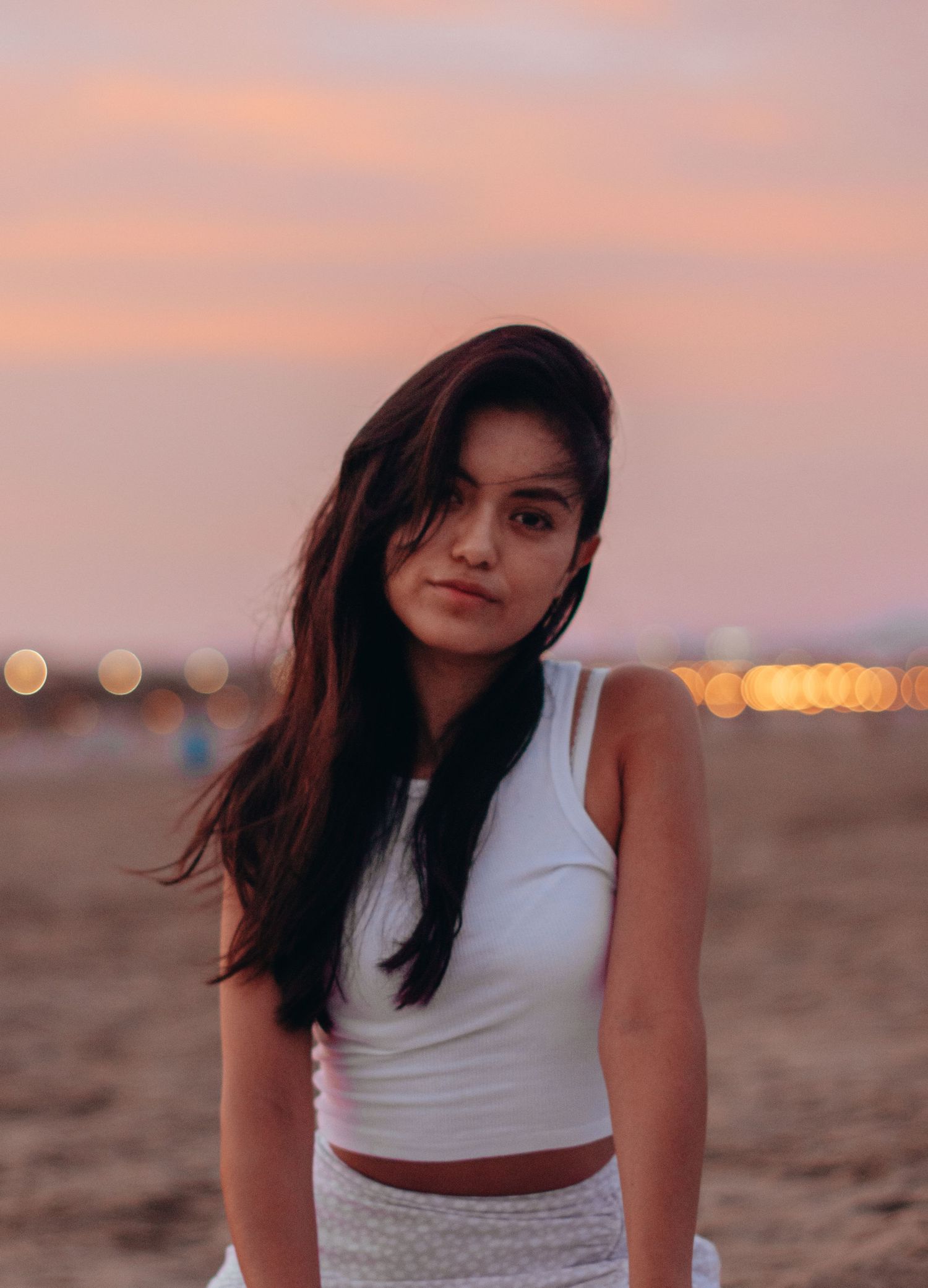 Woman in white outfit poses on beach during sunset with city lights in background.
