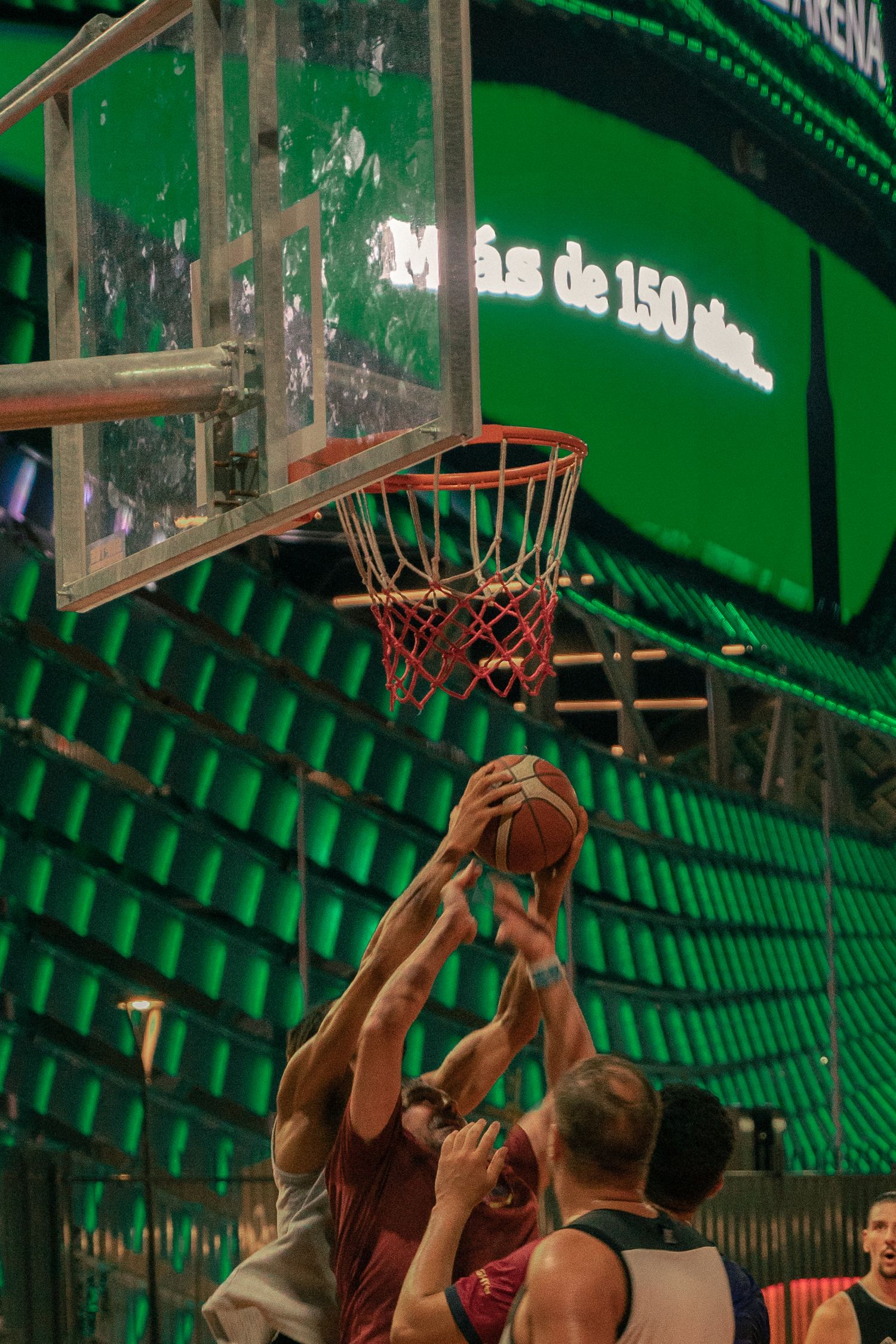 Basketball players jump for a rebound under a glass backboard in a green-lit indoor arena.
