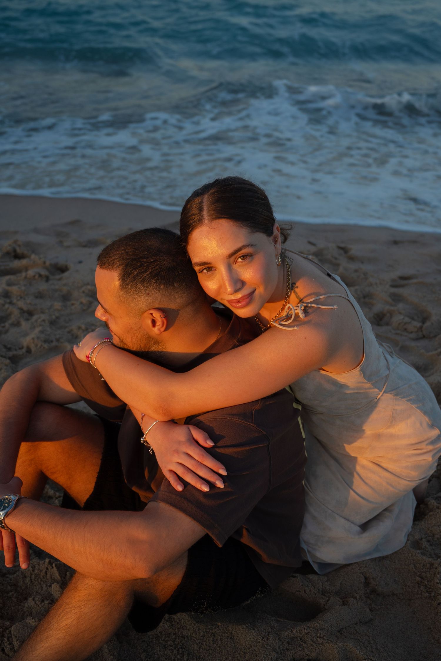A couple embraces on the sandy beach with ocean waves in the background.