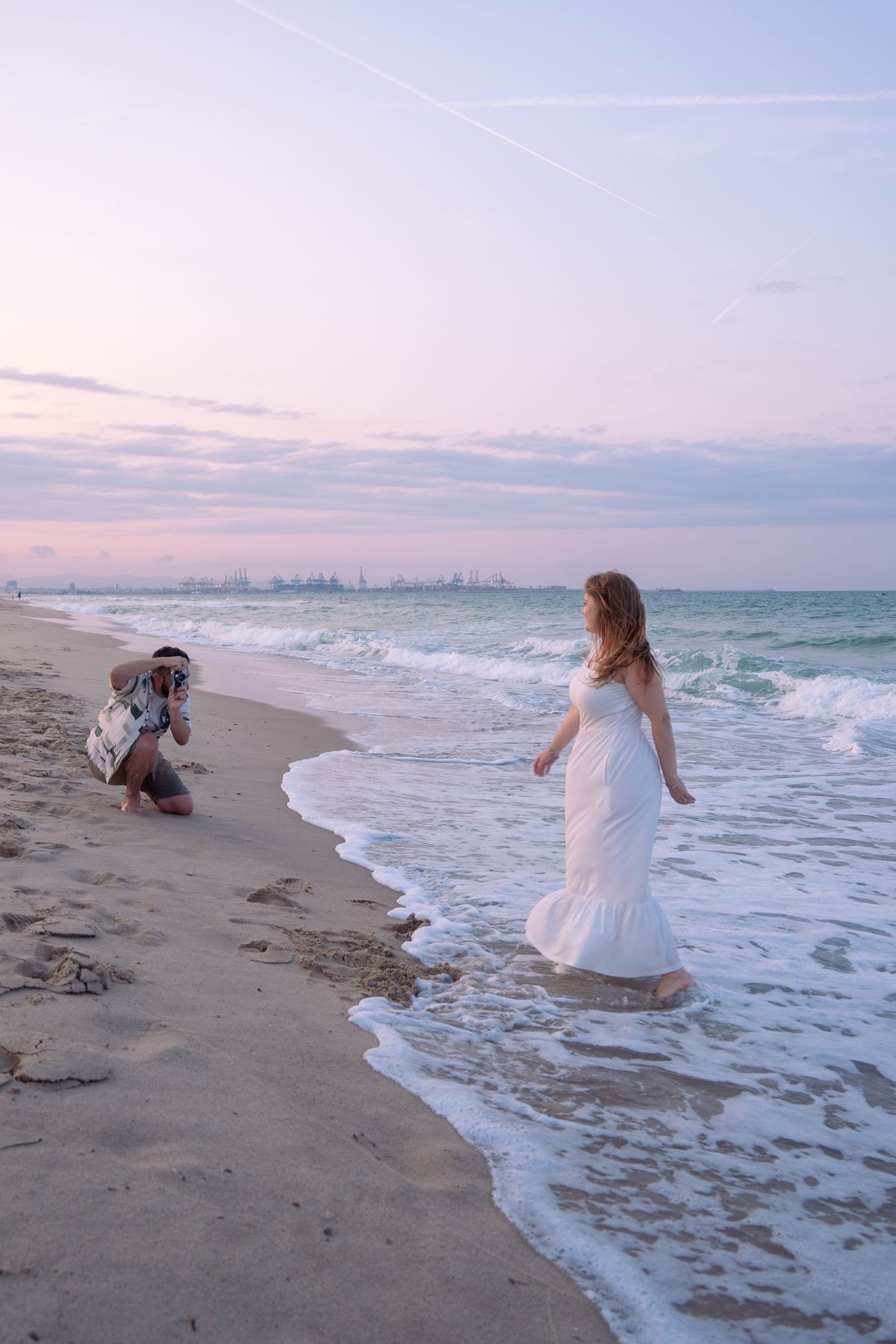 Person in white dress playing in ocean waves at sunset on empty beach.