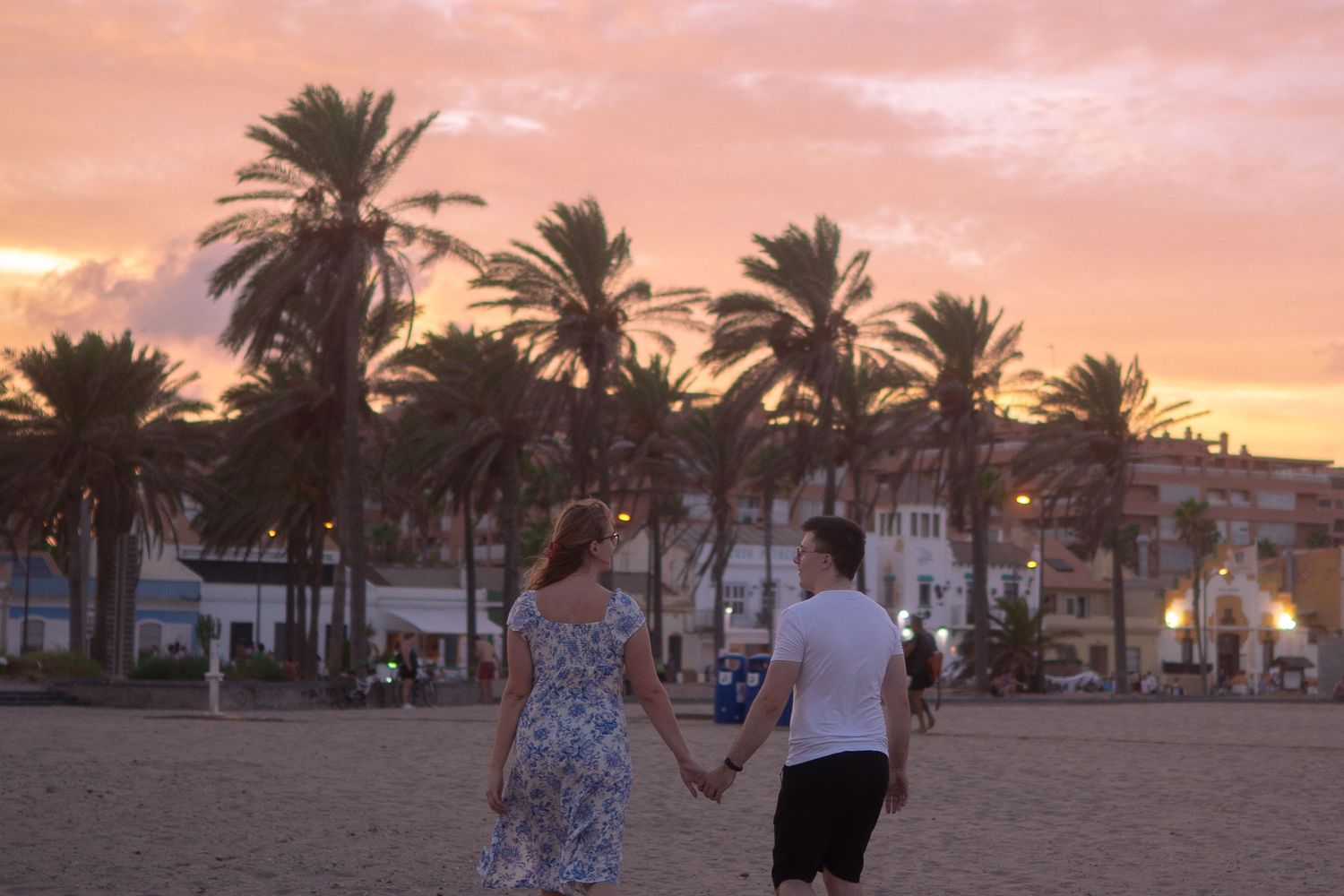 Couple walking hand in hand along a palm-lined beach at sunset.