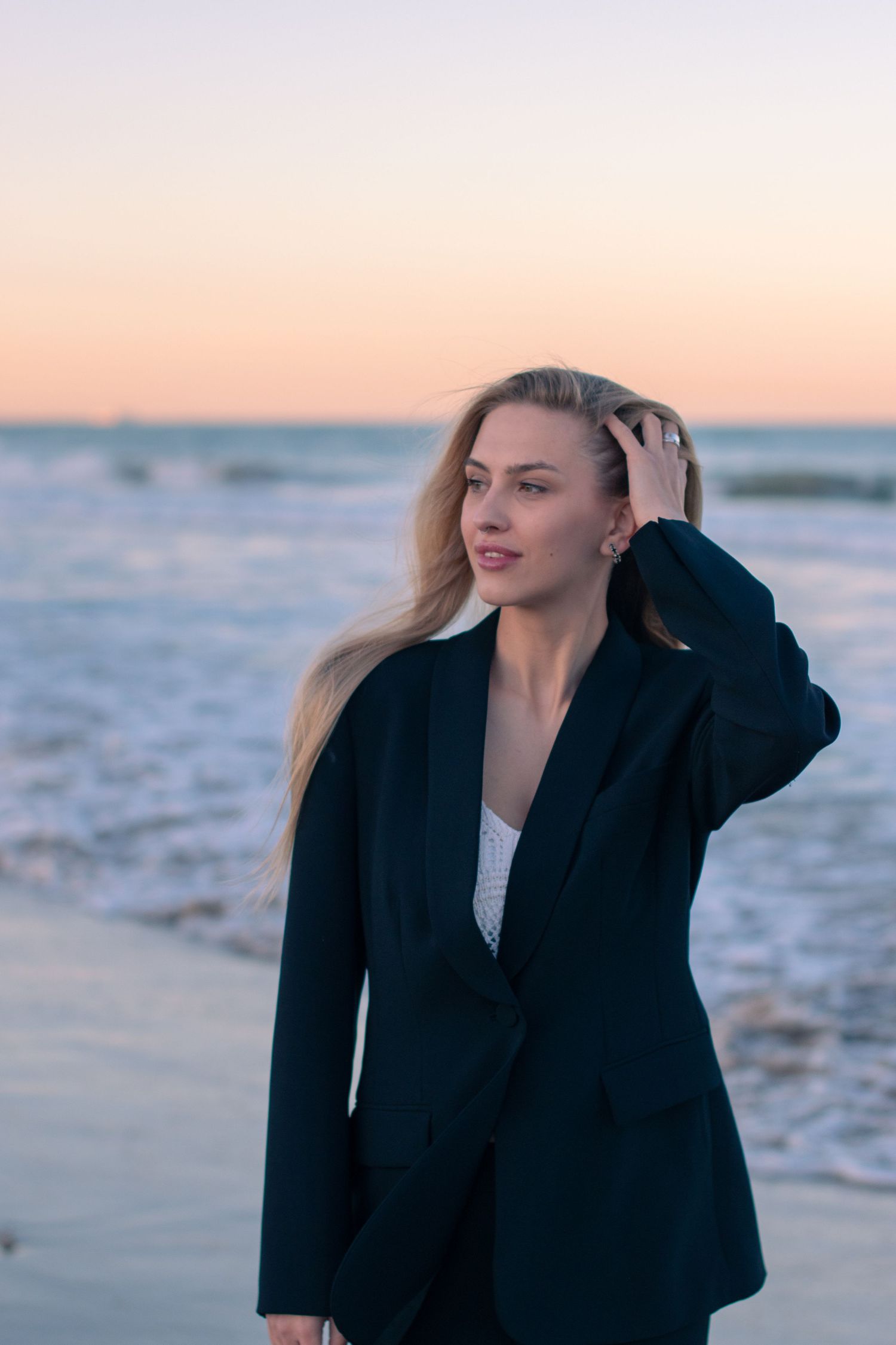 A figure in a black coat poses on the beach at sunset.