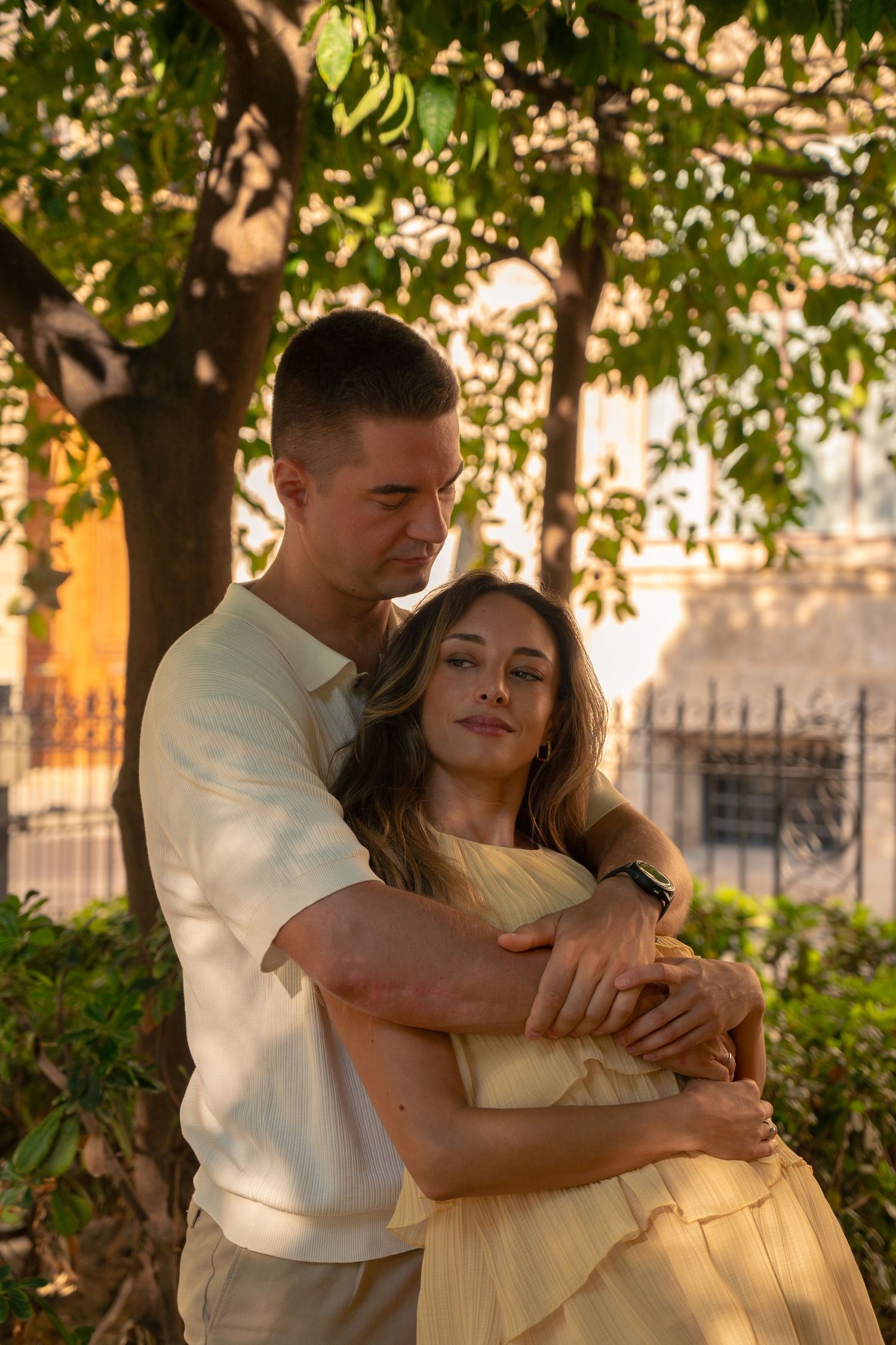 A couple embraces tenderly under a tree at golden hour, the man wrapping his arms around the woman in a yellow dress.