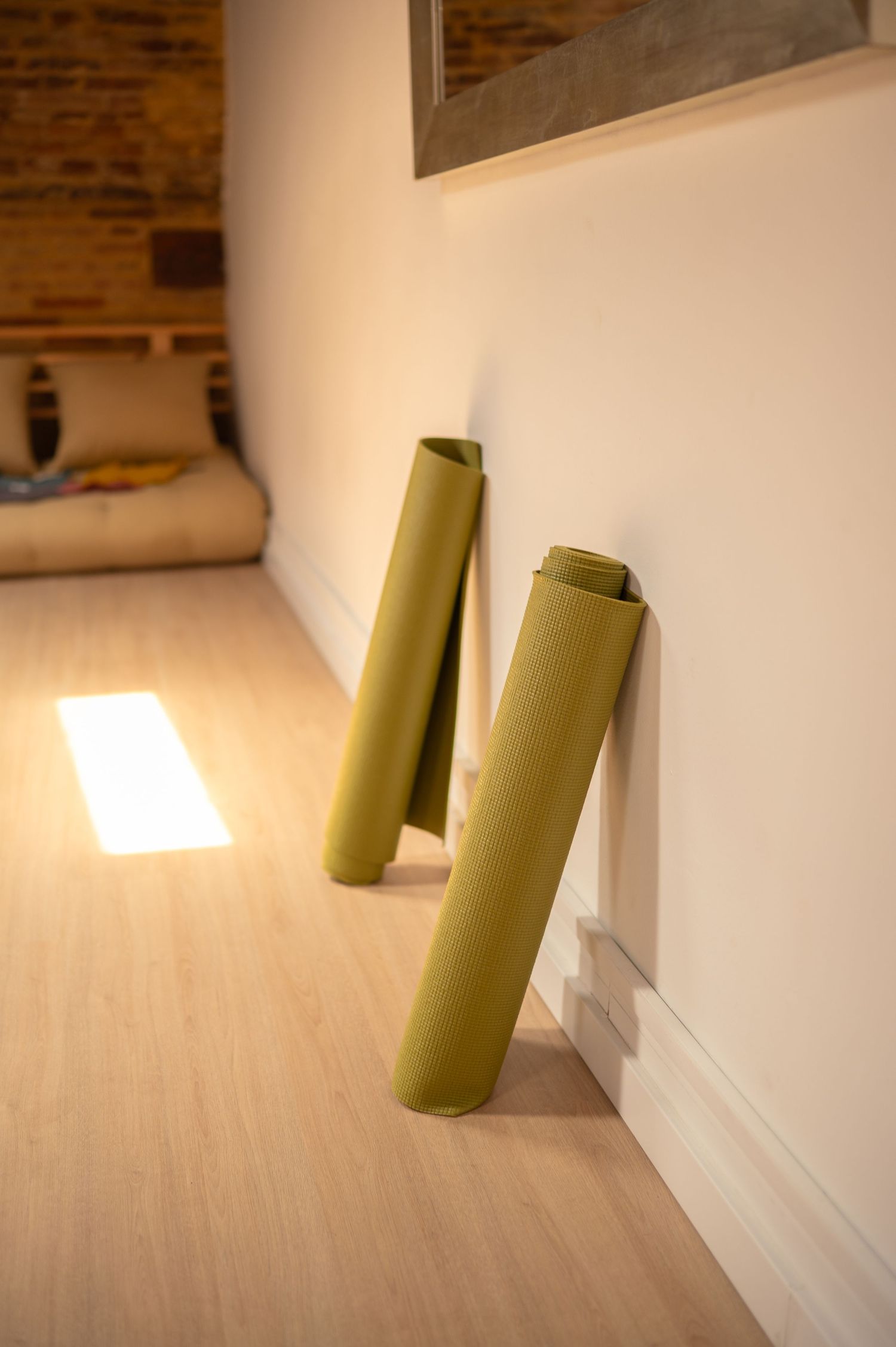 Two olive green yoga mats leaning against a white wall in a sunlit room with wooden floors.