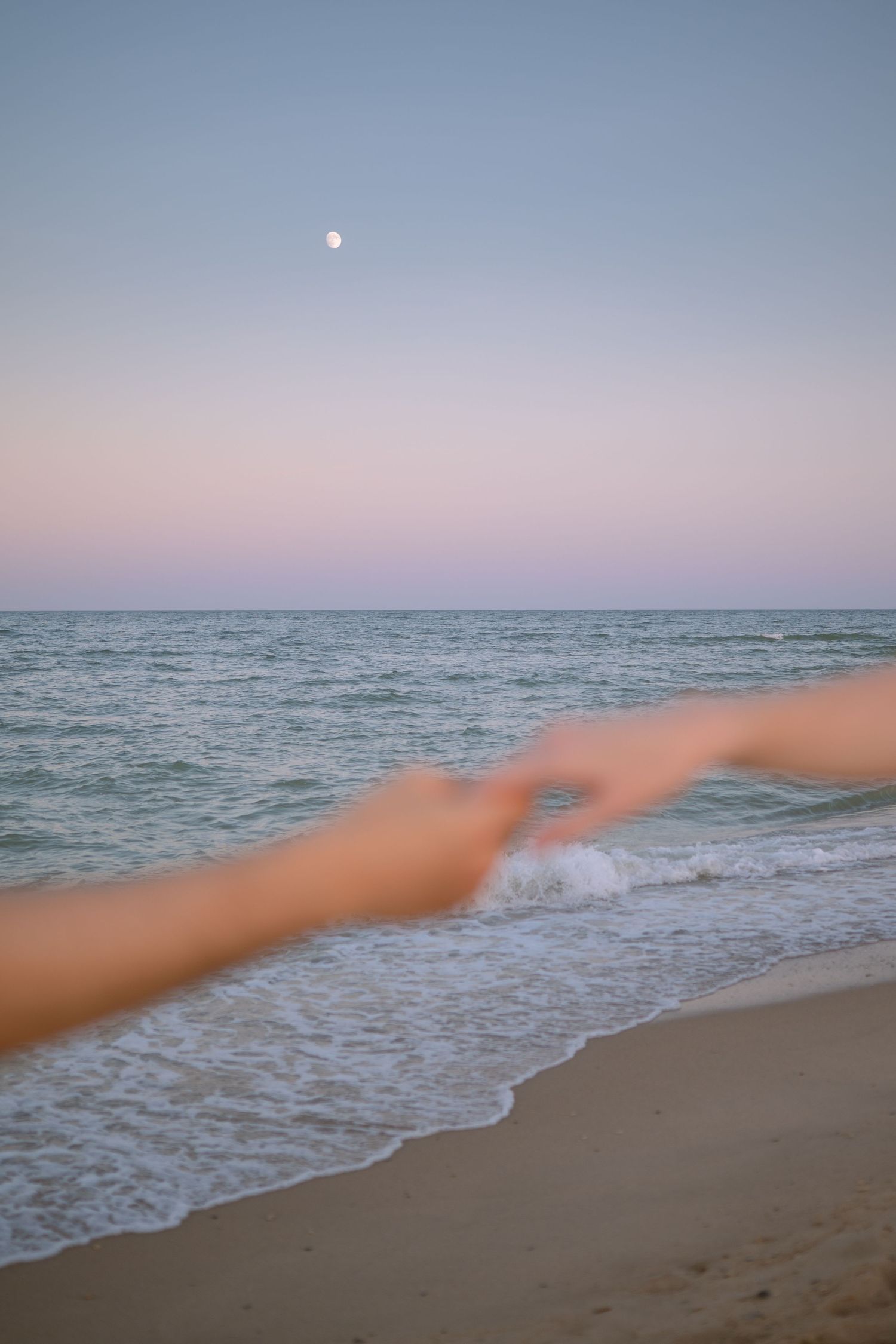 Outstretched arm reaching across a serene beach at sunset with gentle waves rolling in.