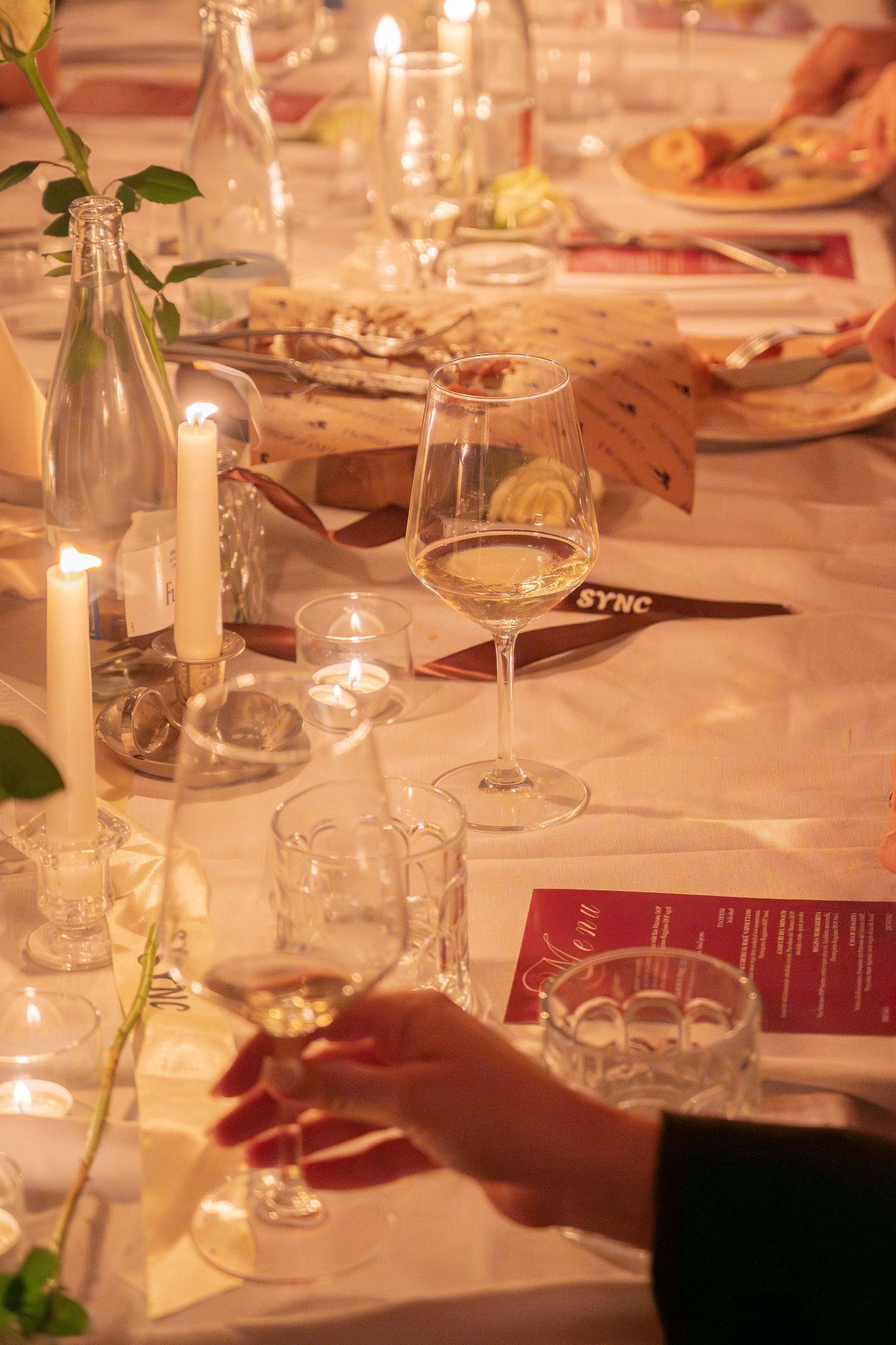 Wine glasses and table settings illuminated by candlelight with menus and decorative greenery.