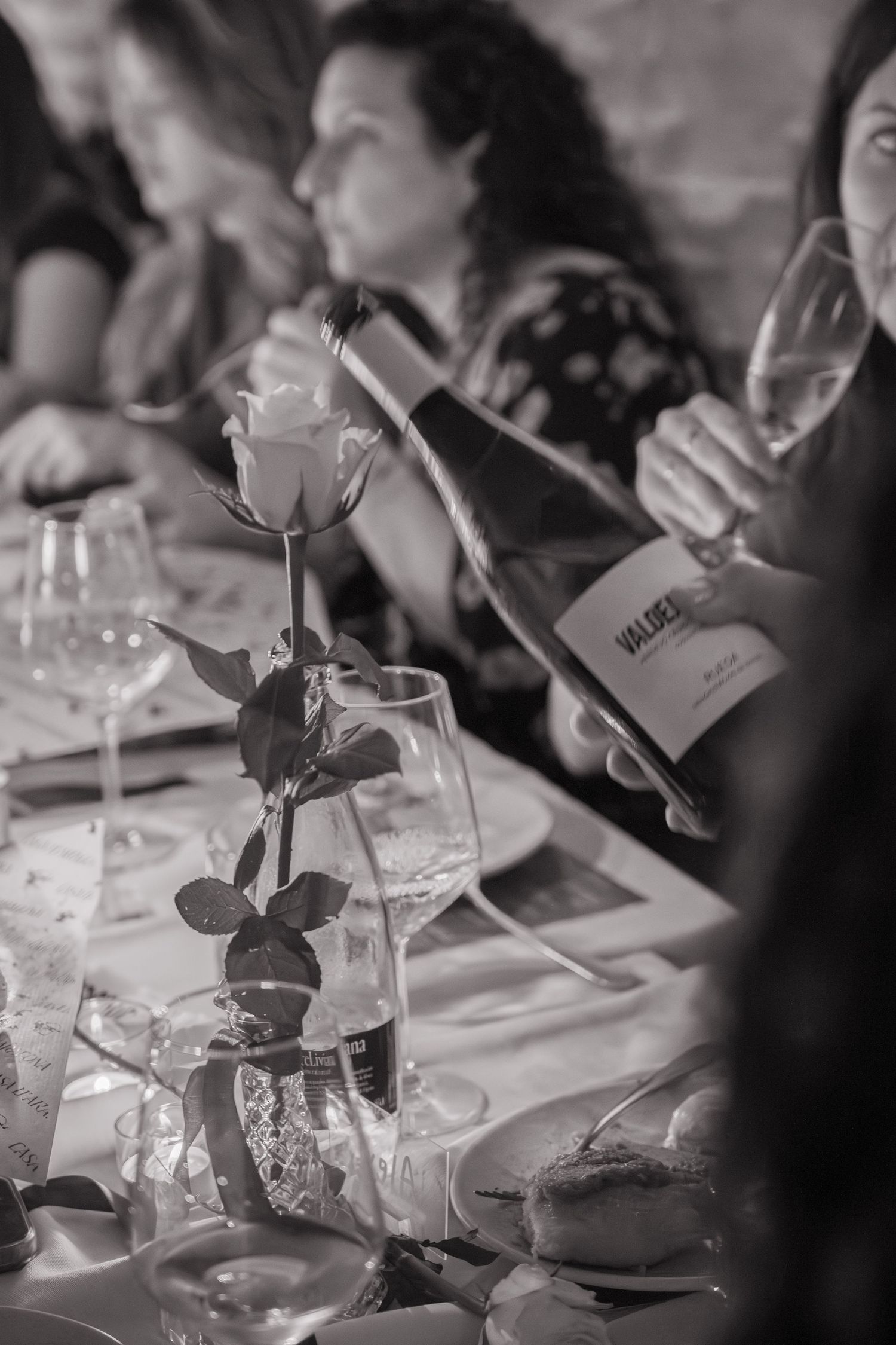 Black and white image of a dinner table with wine bottle and rose in stemmed vase.