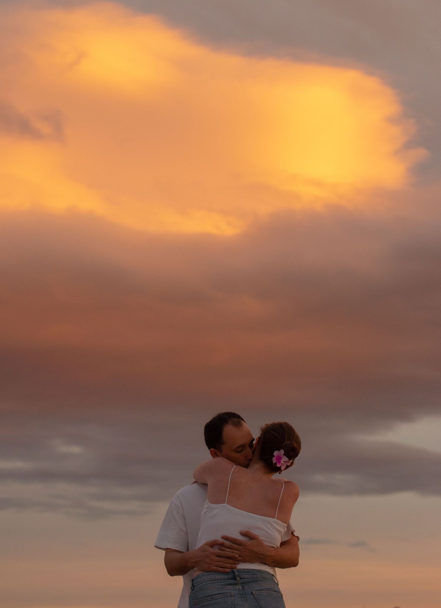 Couple embraces on beach during golden sunset with dramatic orange clouds in the background.