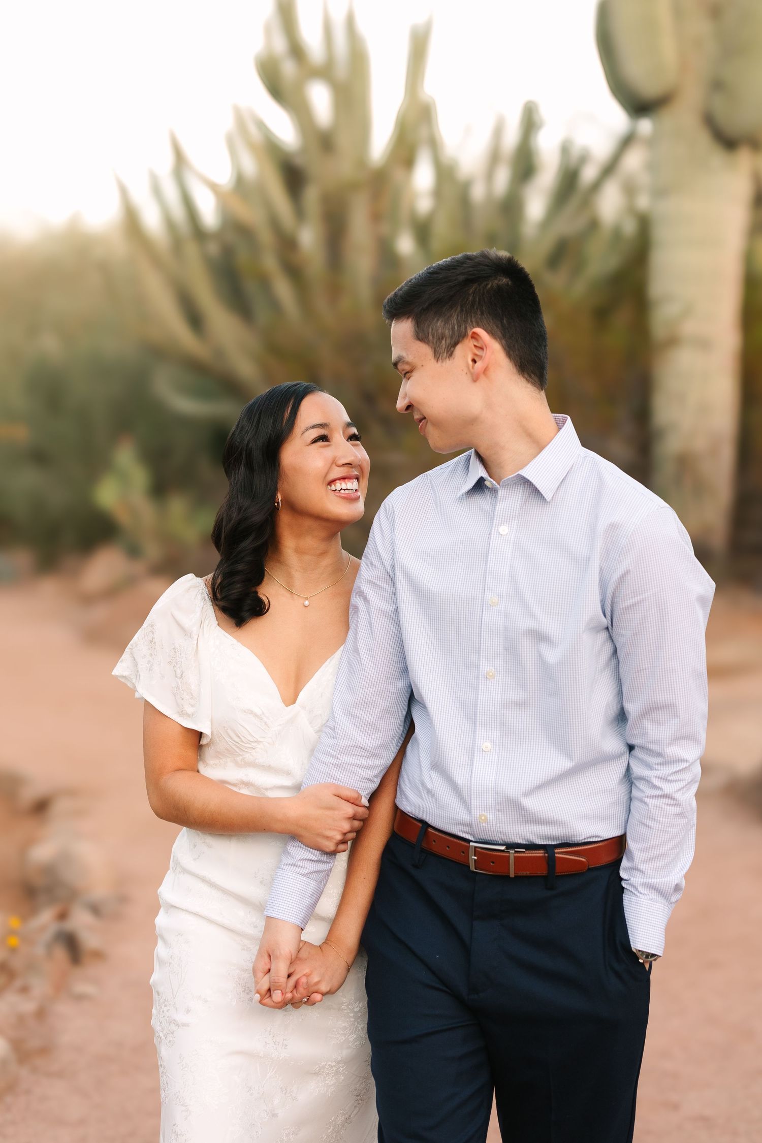 A couple in white and blue attire share tender moments while posing against a desert landscape with cacti.