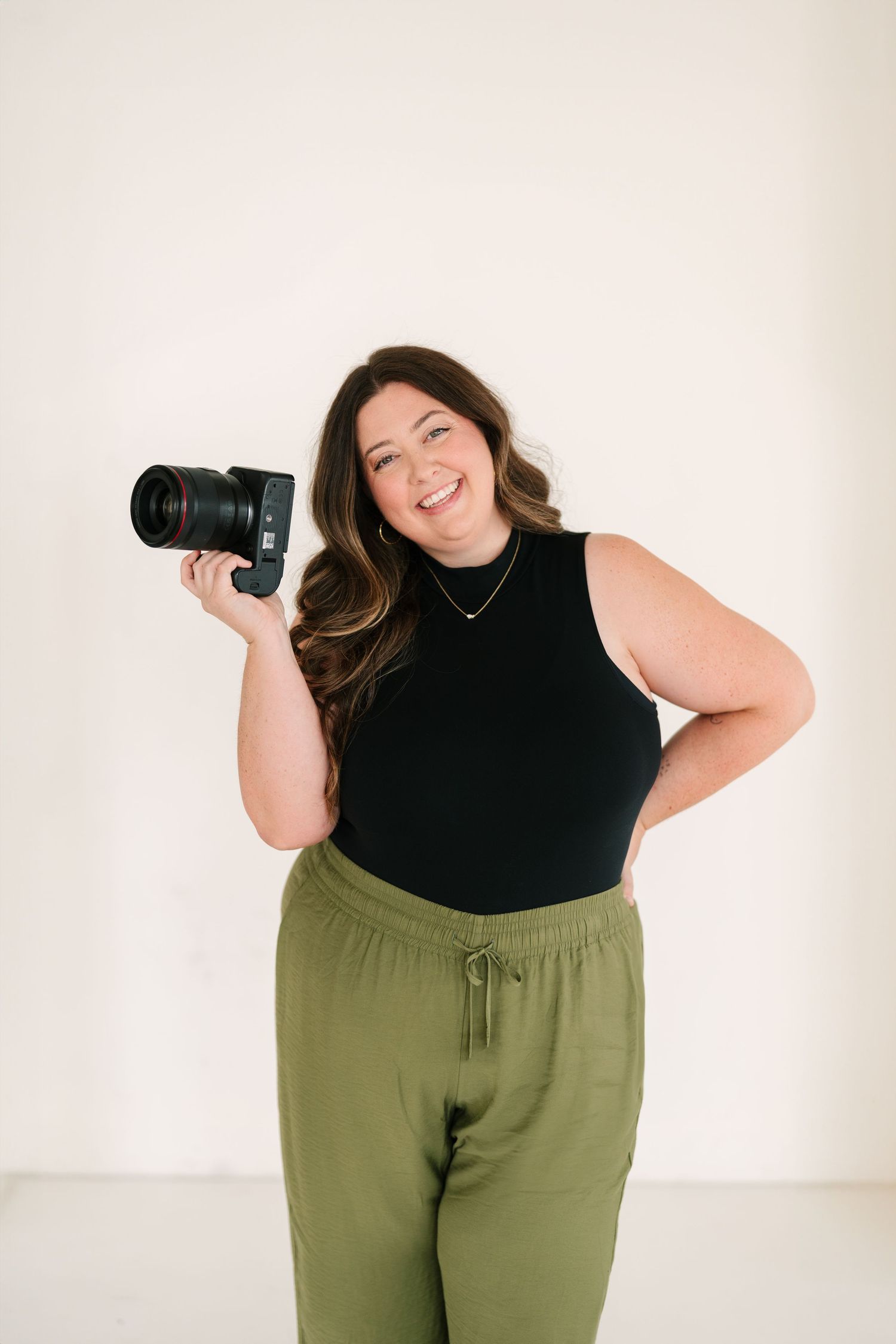 Smiling female photographer holding a DSLR camera, wearing black top and olive green pants against white background.