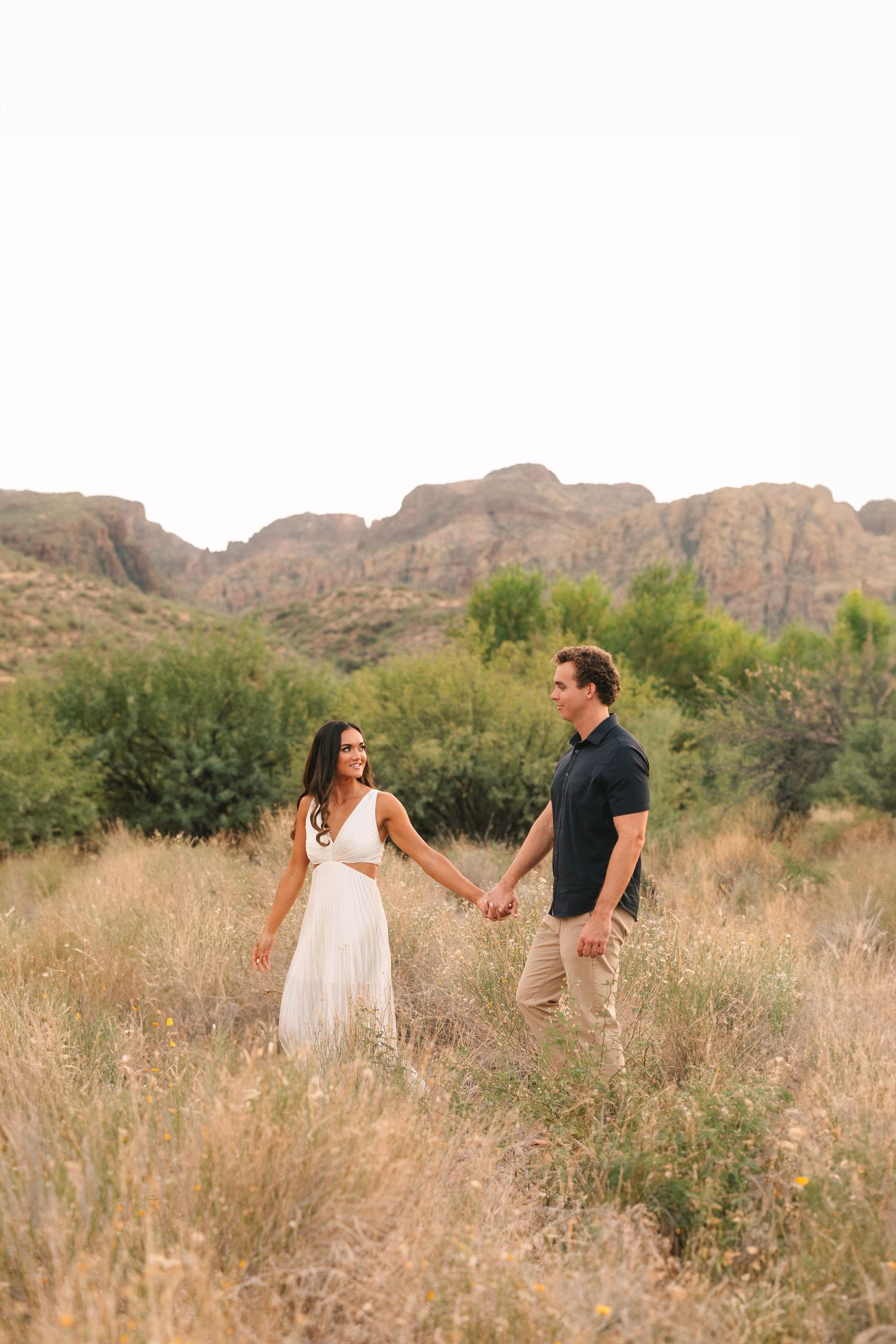 A couple walks and holds hands through a sun-drenched grassy field with majestic desert mountains in the background.