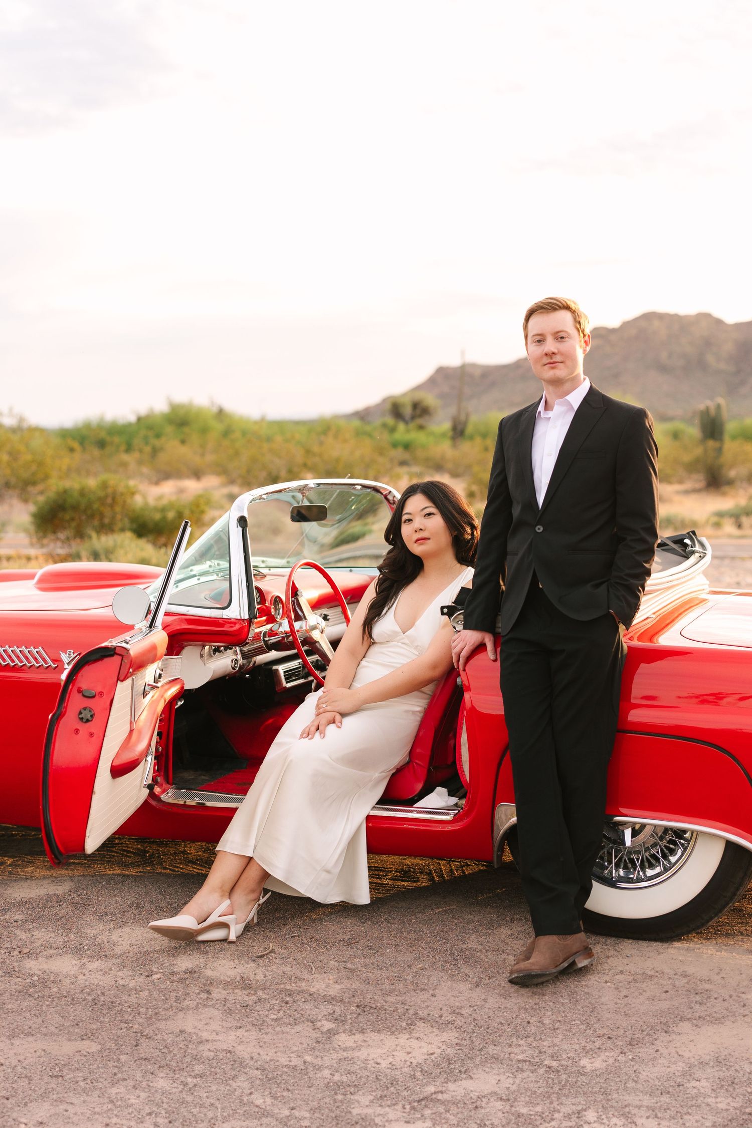 A couple poses elegantly with a classic red Thunderbird car in a desert setting at sunset.