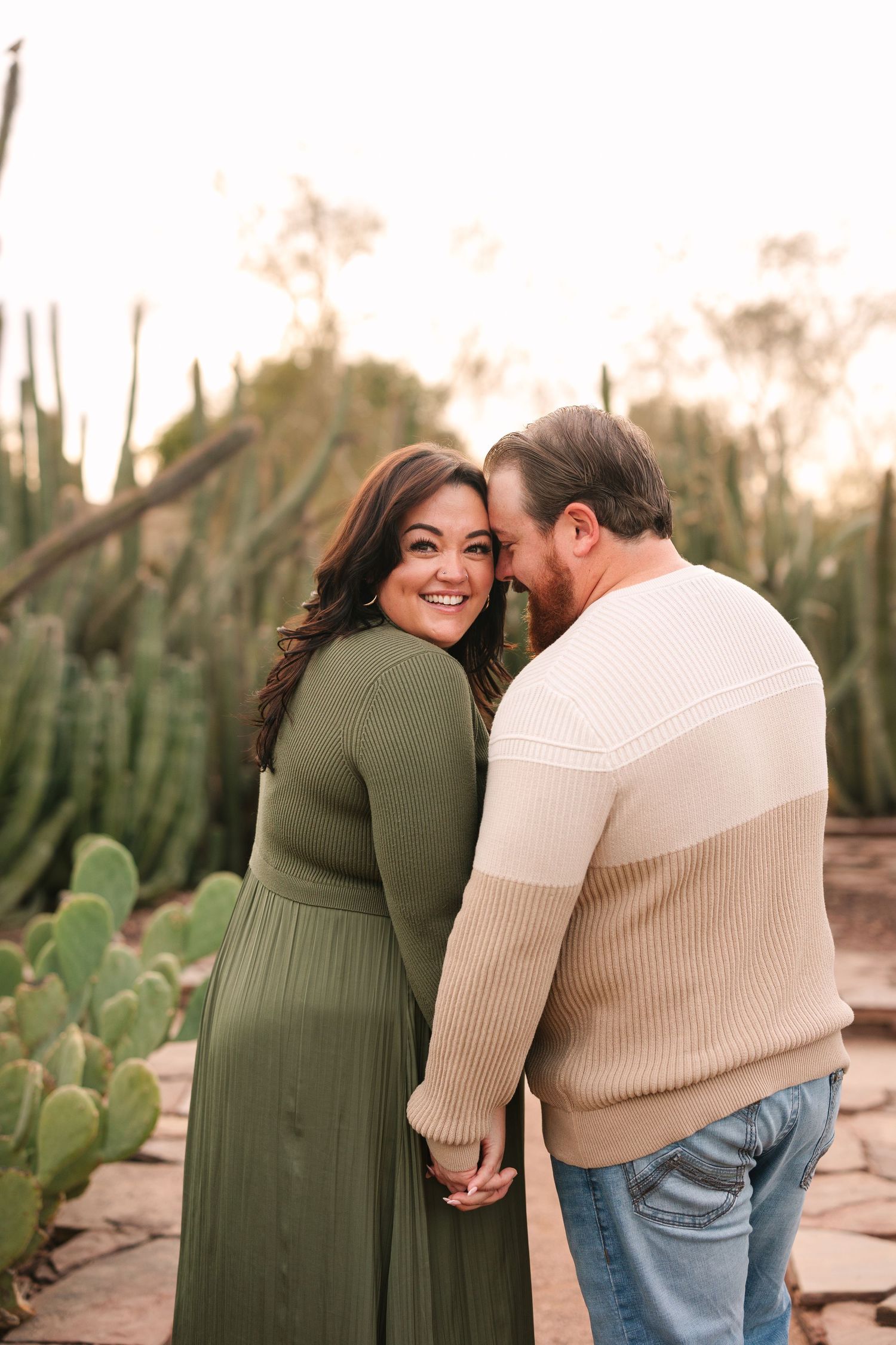 A couple shares tender moments against a desert landscape with cacti at sunset, wearing green dress and beige sweater.