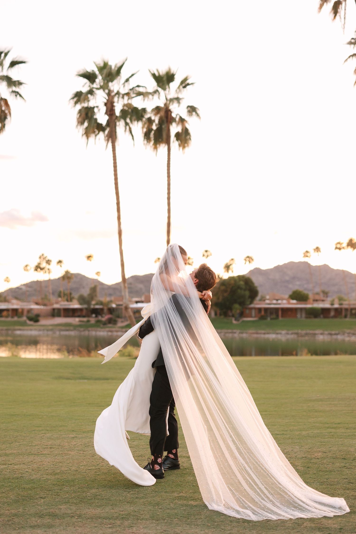 A couple embraces on a golf course at sunset with palm trees and mountains in the background, flowing veil catching the breeze.