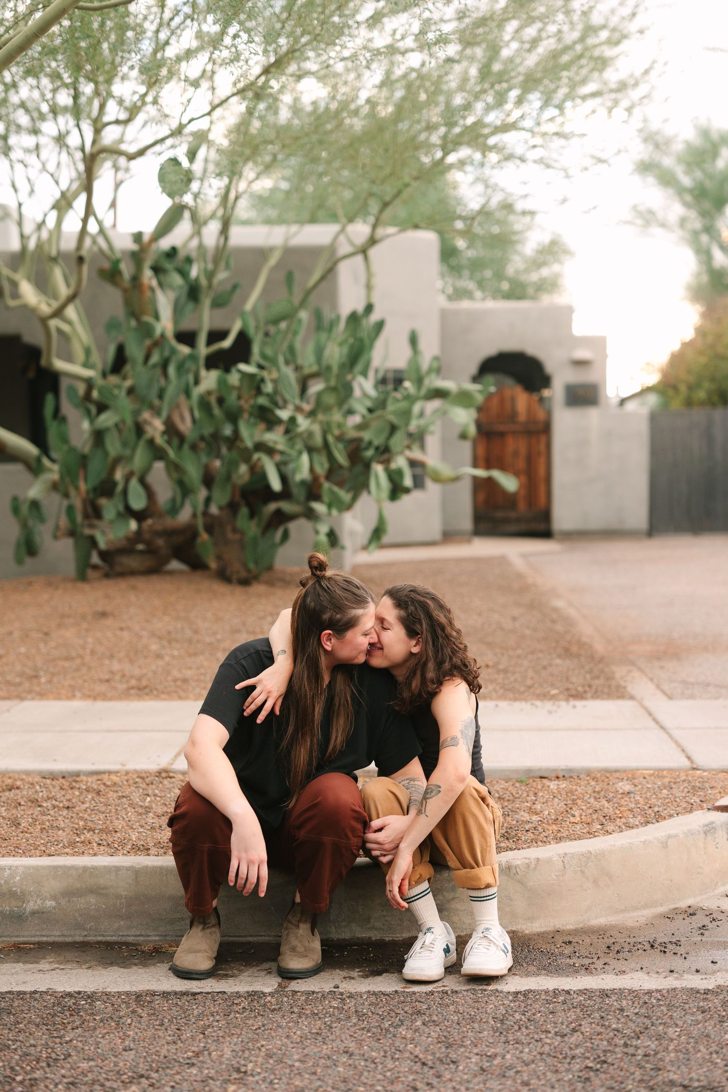 Romantic street photoshoot featuring a couple sitting and embracing on paved sidewalk beside desert landscaping.