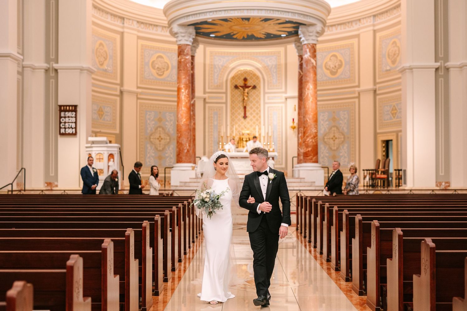 Couple walks down the aisle of an elegant church with cream-colored walls and ornate details.