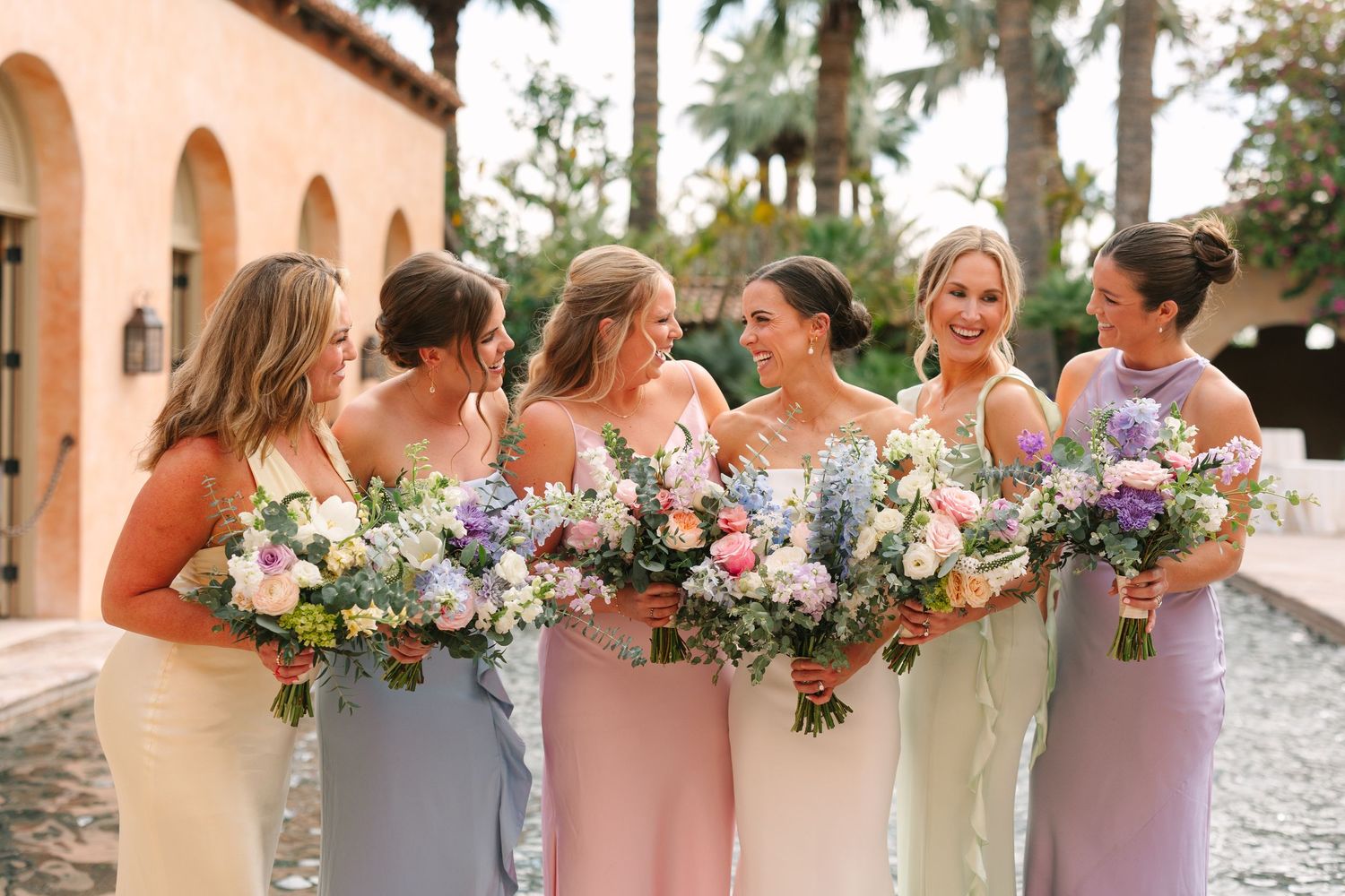 Bridesmaids in pink dresses pose with colorful bouquets against stone archway.