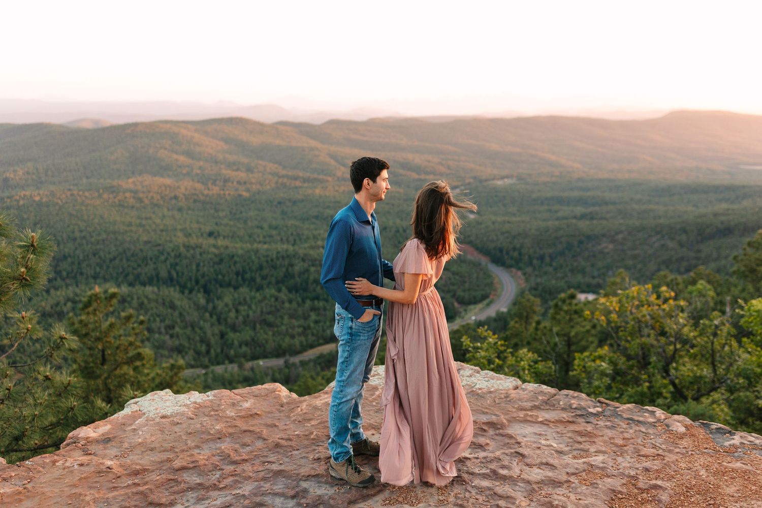 A couple stands atop a mountain overlook during golden hour with rolling forested hills stretching into the distance.