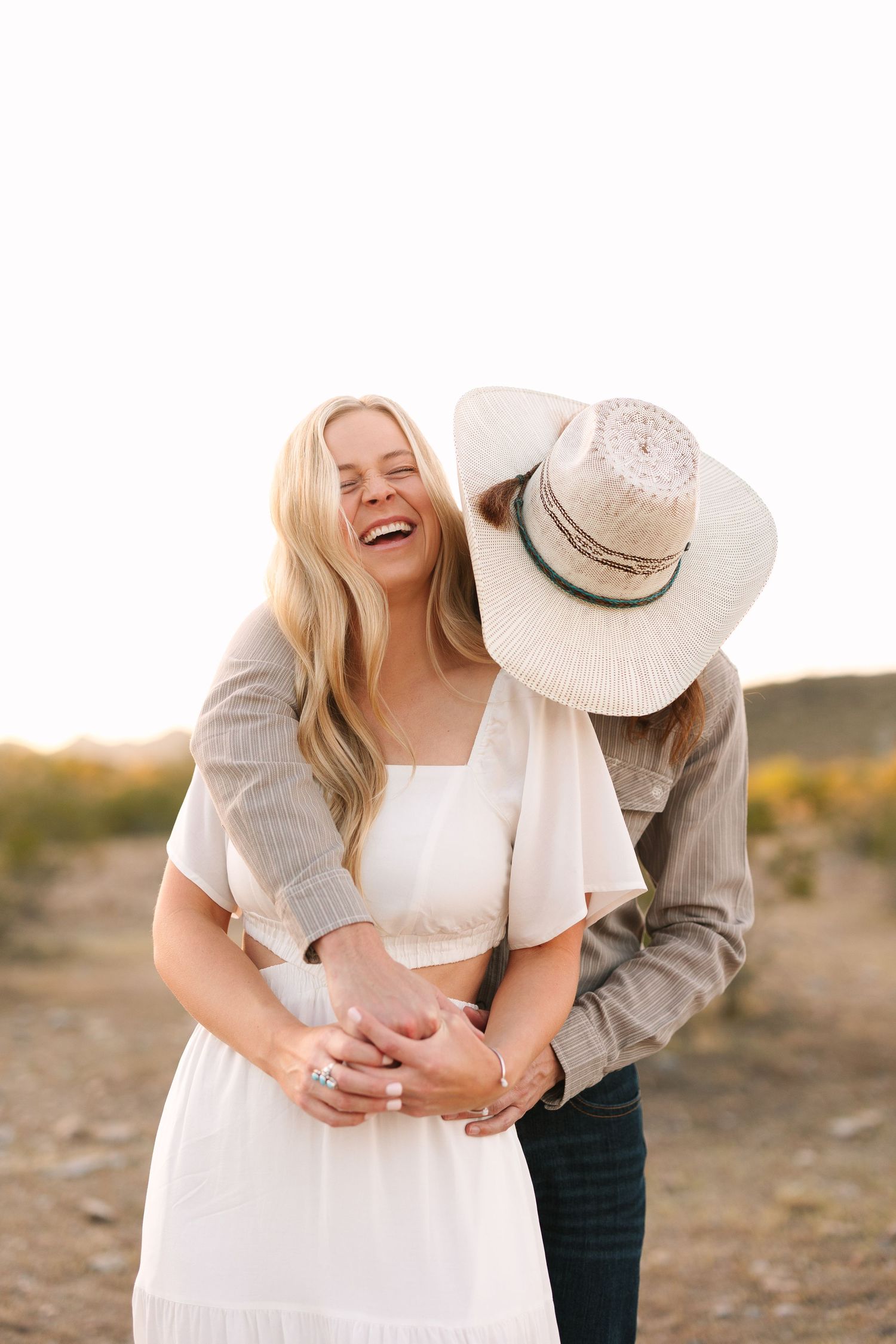 A couple shares a joyful moment in a desert landscape at sunset, with the person wearing a cowboy hat embracing from behind.
