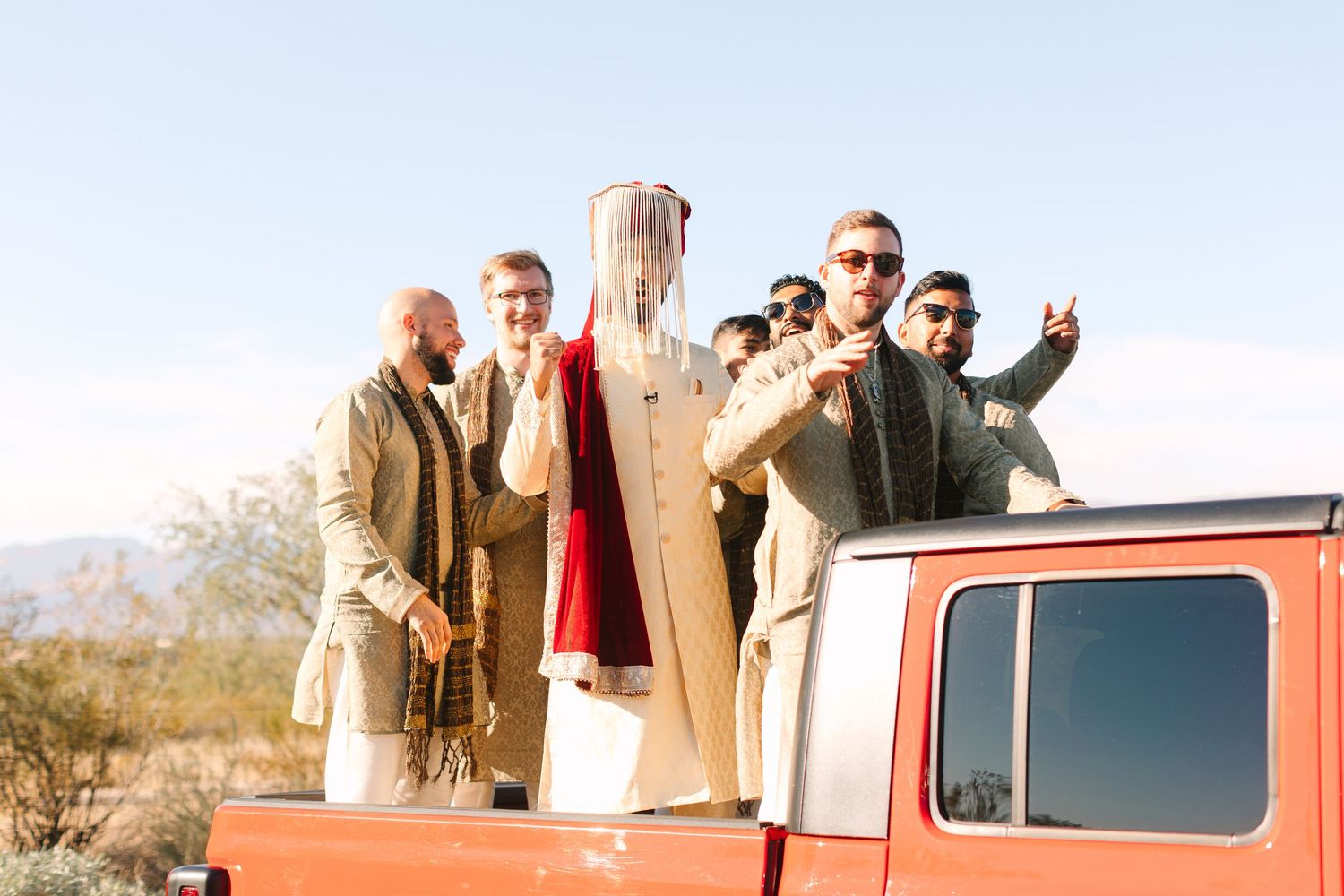 Group poses on vintage orange truck in desert setting with mountains in background.