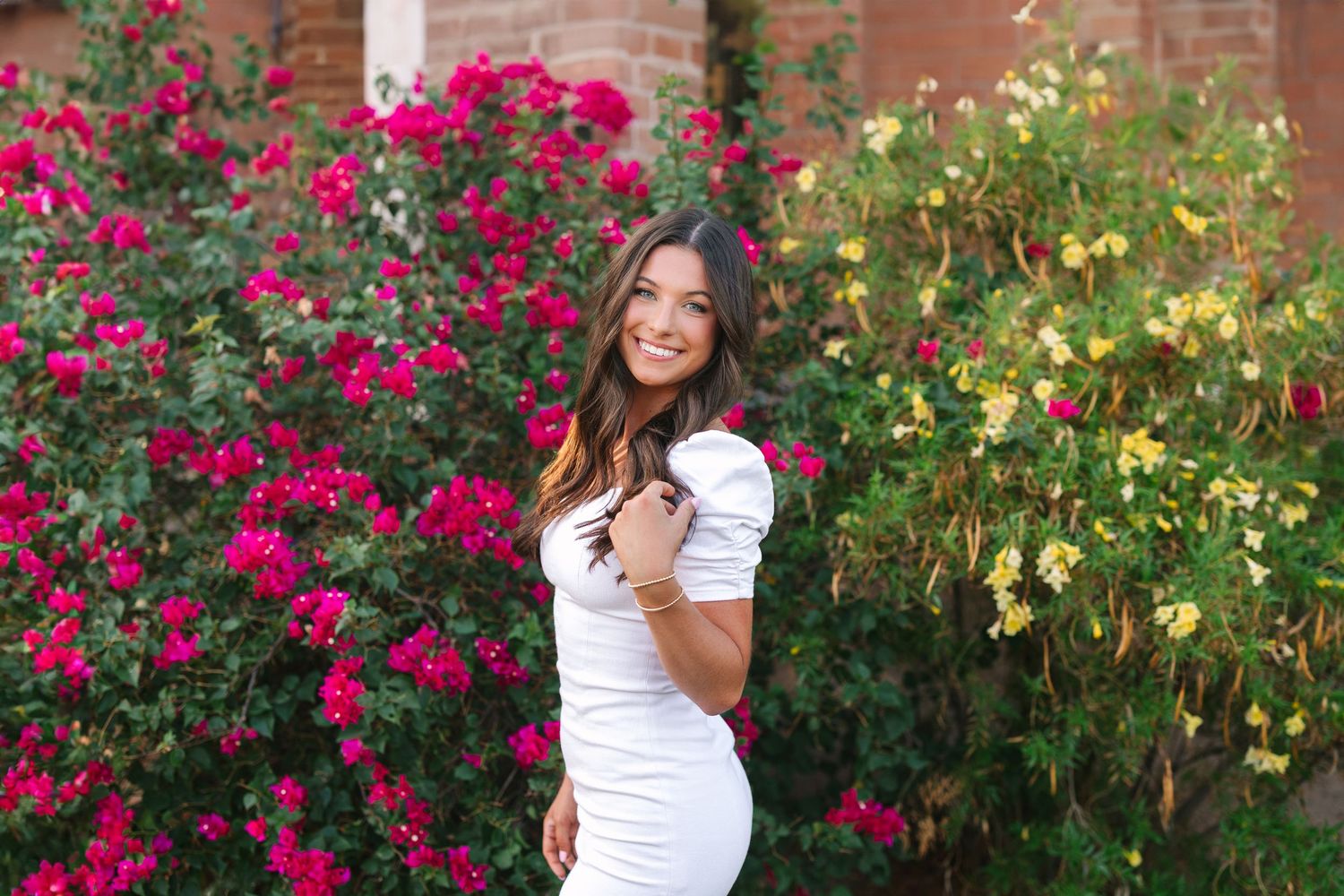 A person in a white top poses in front of vibrant pink rose bushes and yellow flowers in a garden setting.