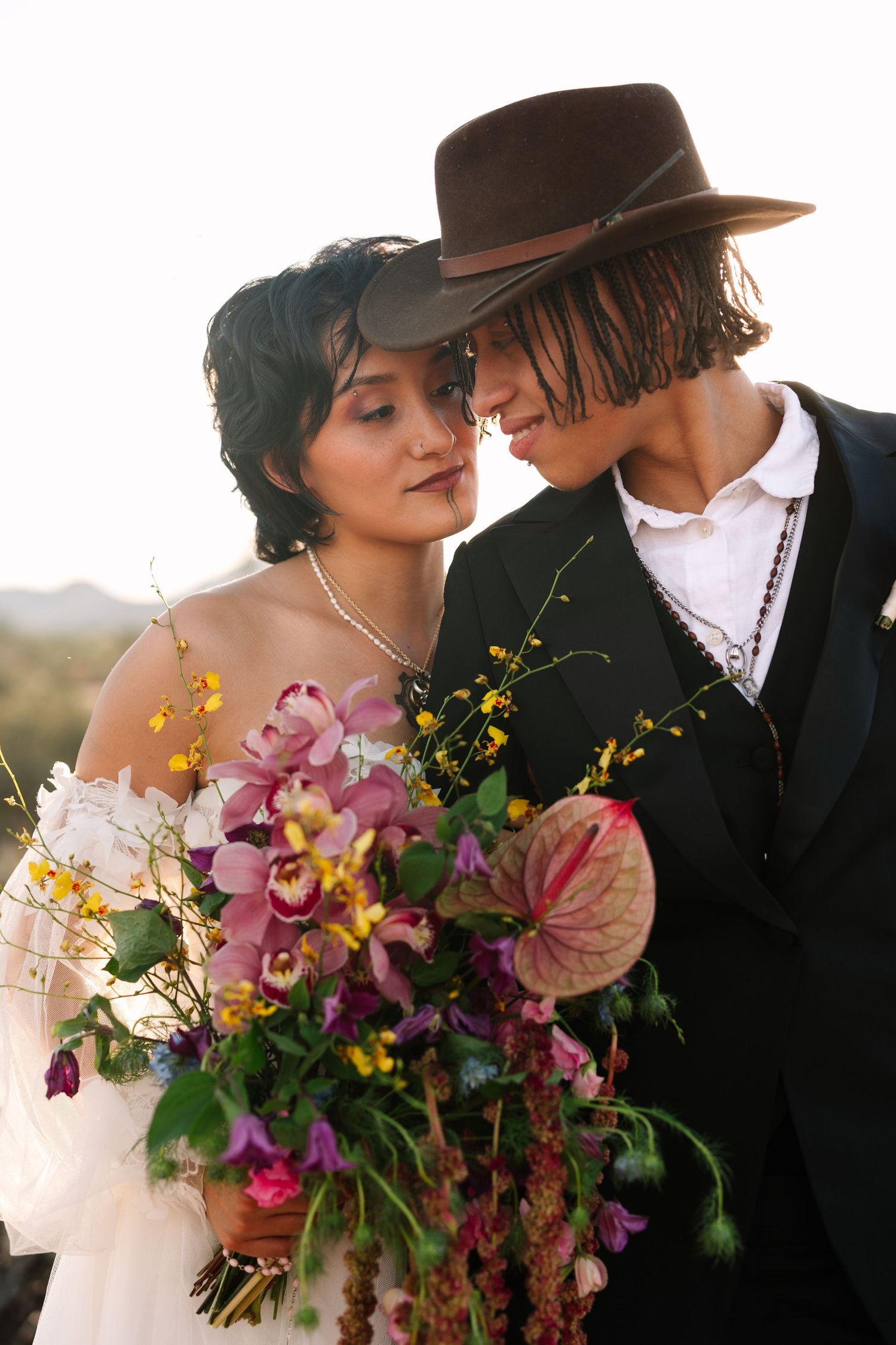 A bride and groom share an intimate moment, she holds a vibrant bouquet of orchids and tropical flowers, he wears a brown wide-brim hat.