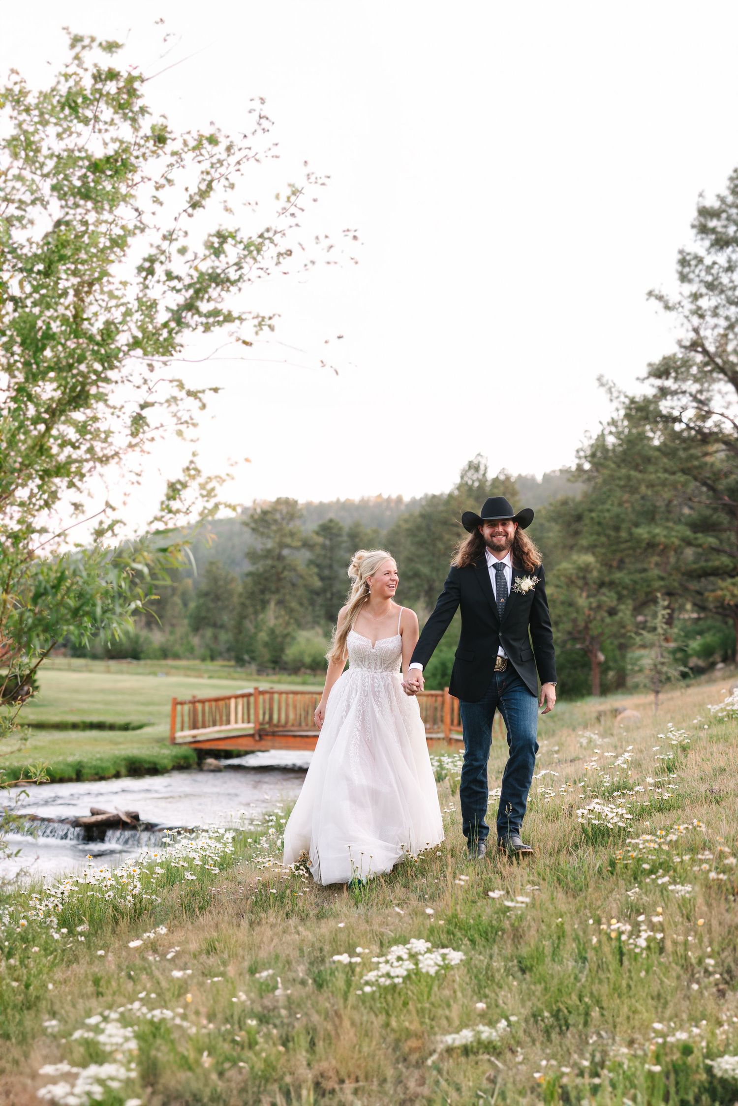 Rustic countryside wedding scene with couple walking through wildflower field near wooden bridge.