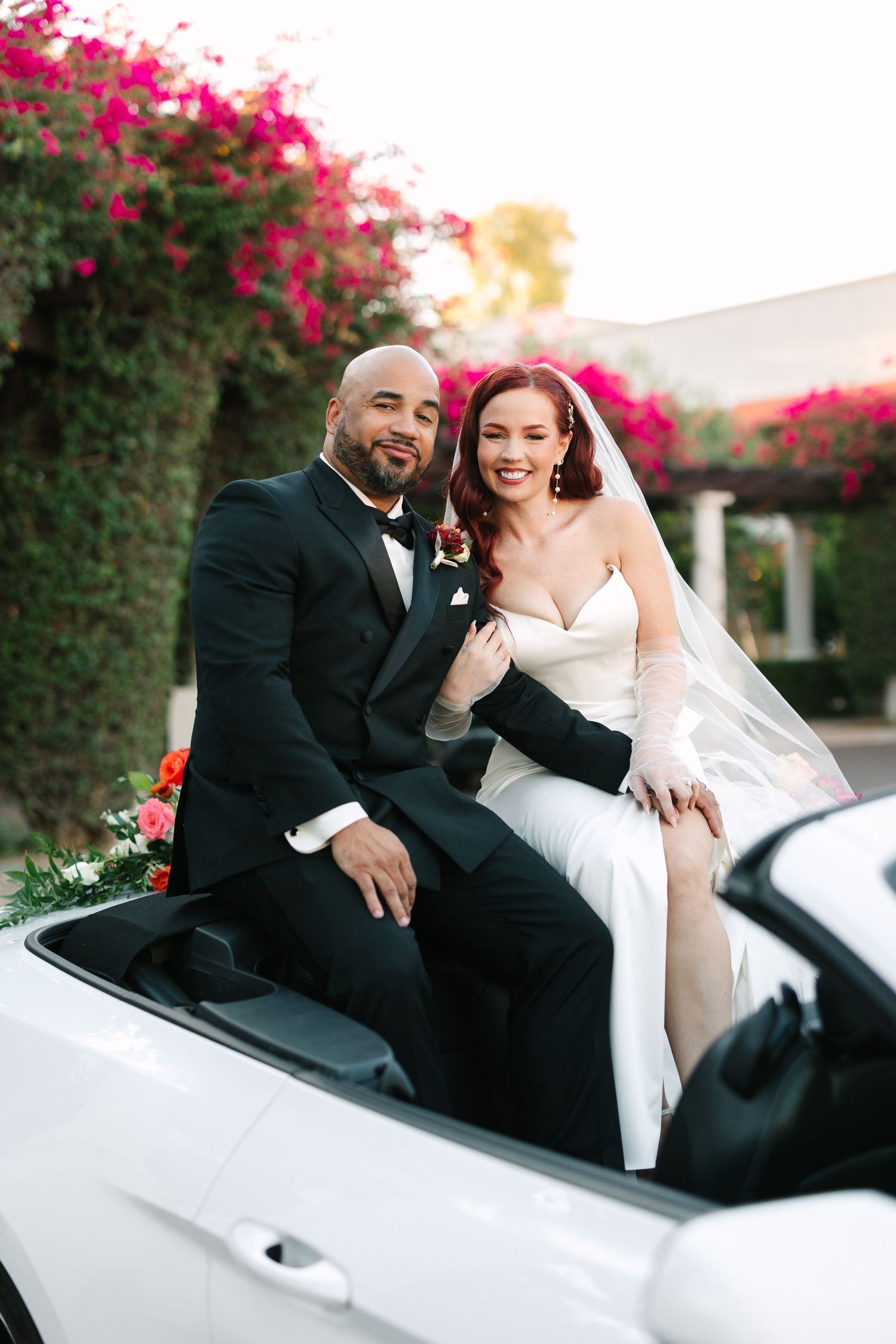 Wedding couple shares intimate moments while sitting in a white vintage car surrounded by pink flowers. Molly McPhoto