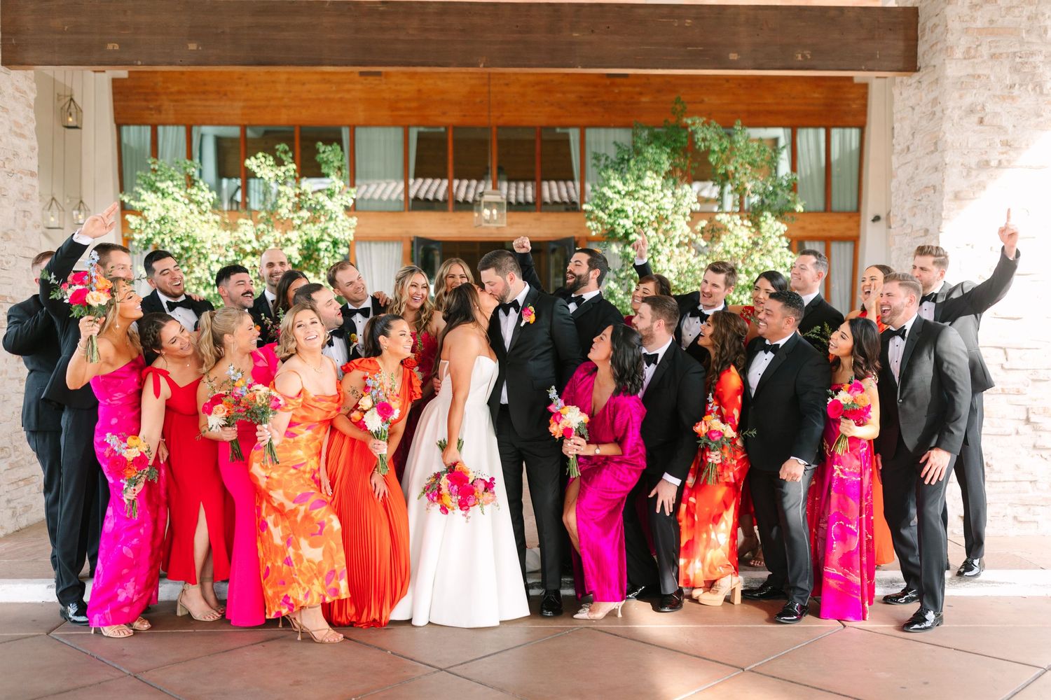 Wedding party in vibrant red and pink attire poses together under covered walkway with floral arrangements.