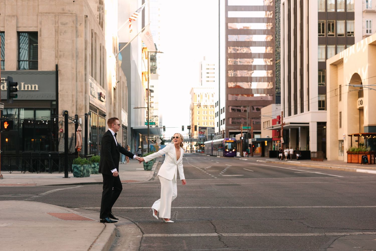 A bride and groom dance playfully together on empty city streets between tall urban buildings.
