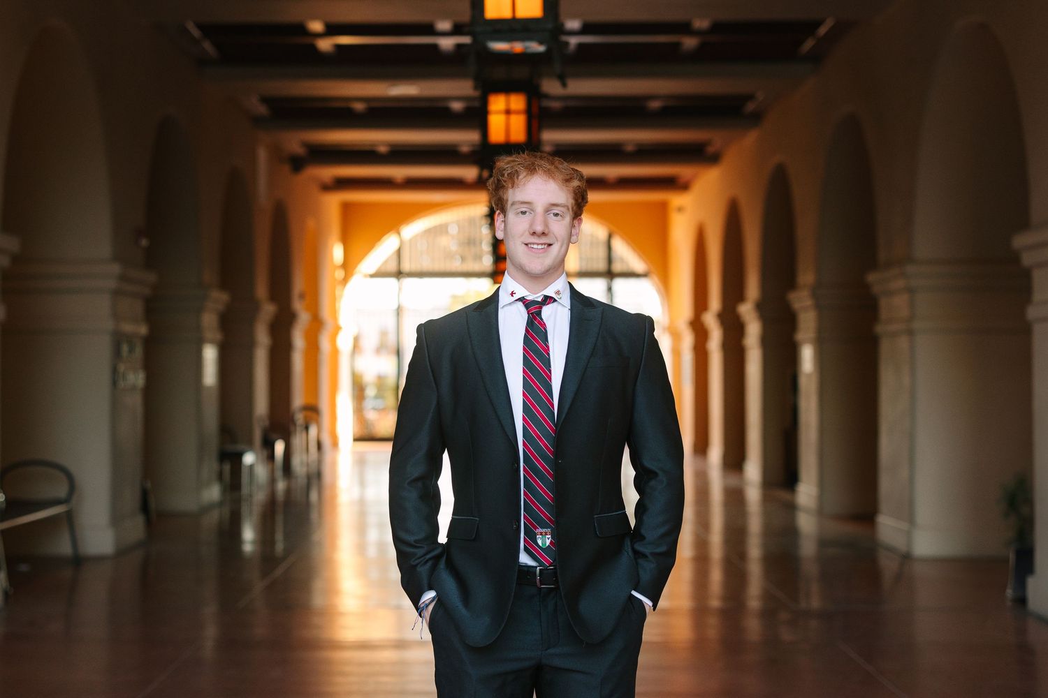 Professional in a dark suit and burgundy tie stands in an elegant arched corridor with dramatic lighting.