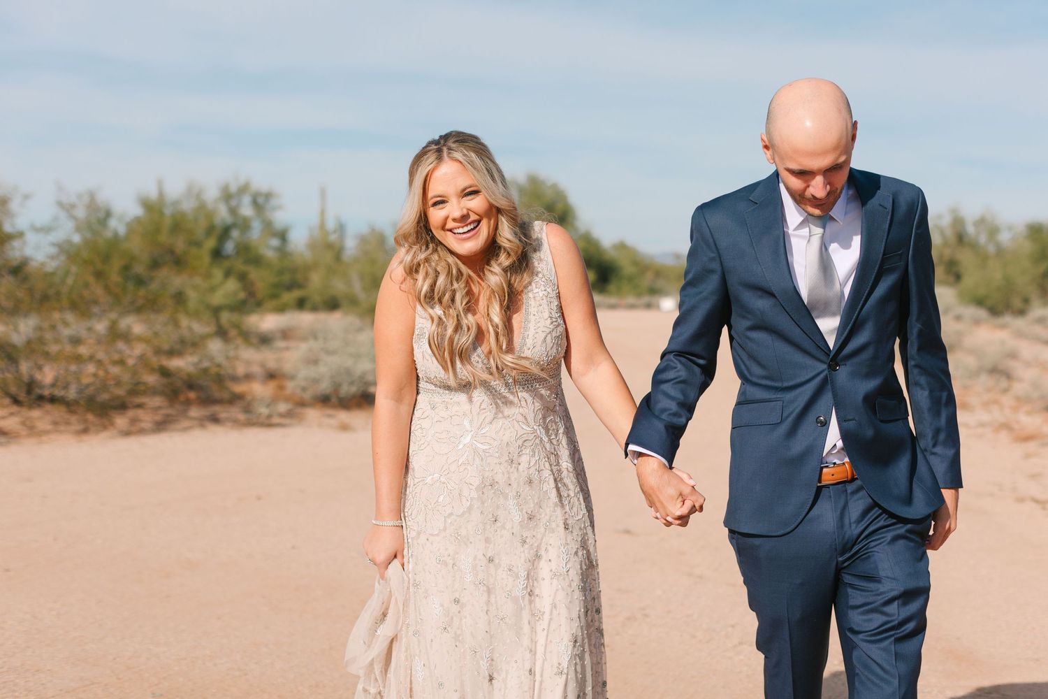 Couple walking hand in hand along a sandy beach in formal wedding attire.