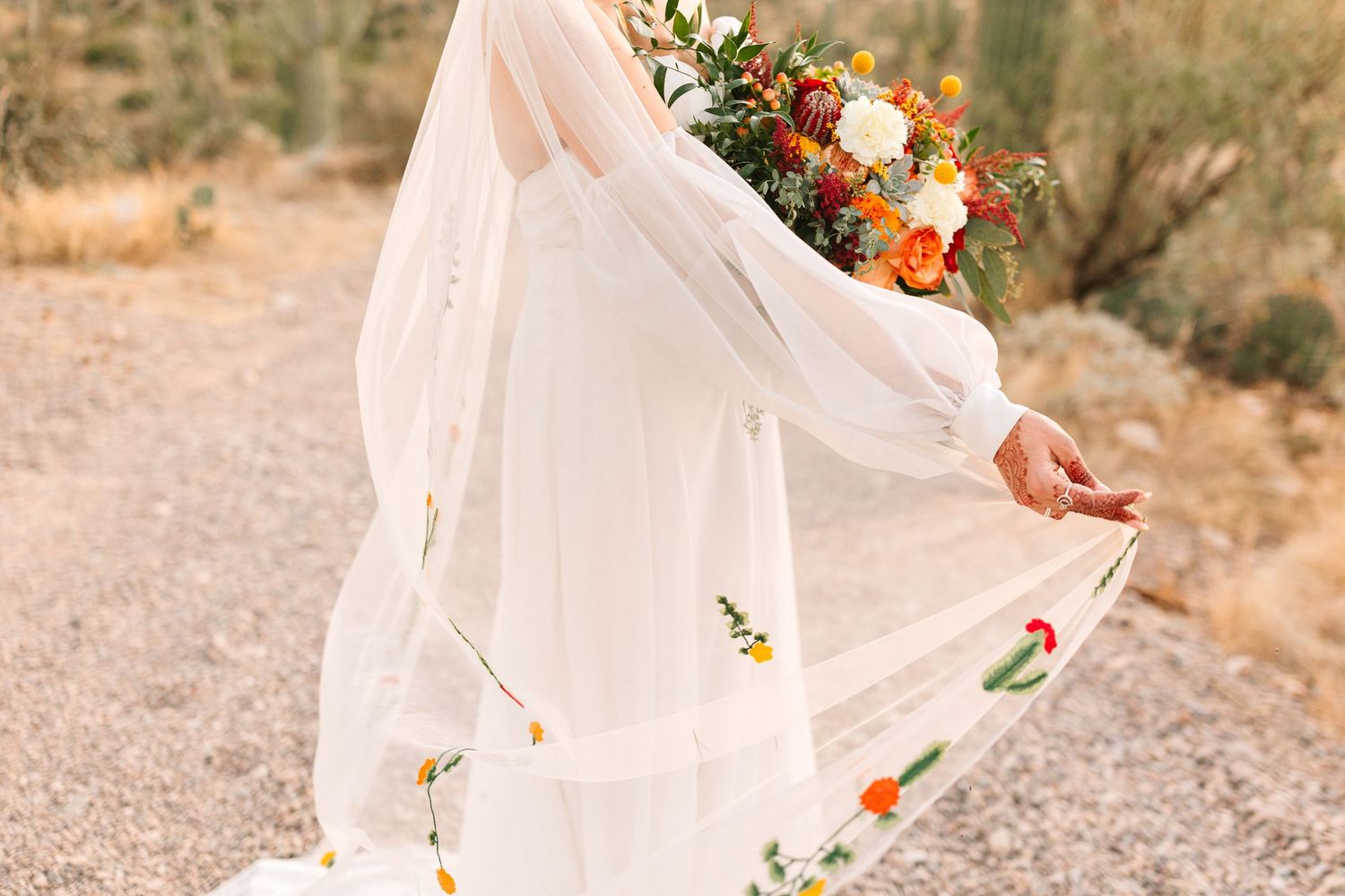 White flowy wedding dress with embroidered flowers twirls in a desert setting with orange and white floral bouquet.