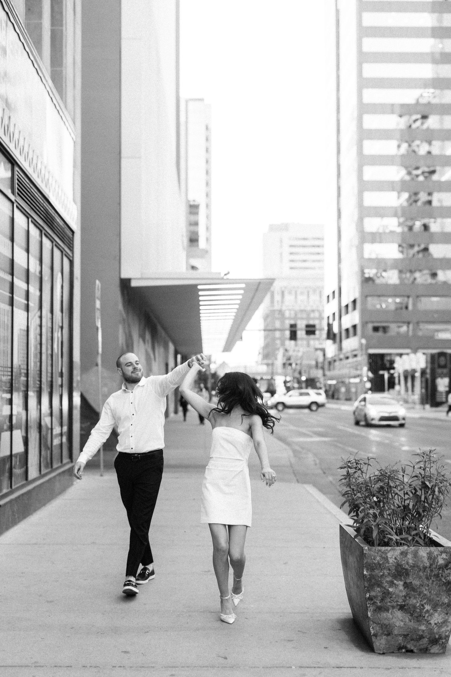 A couple dances joyfully together on an urban sidewalk in front of modern buildings in black and white photography.
