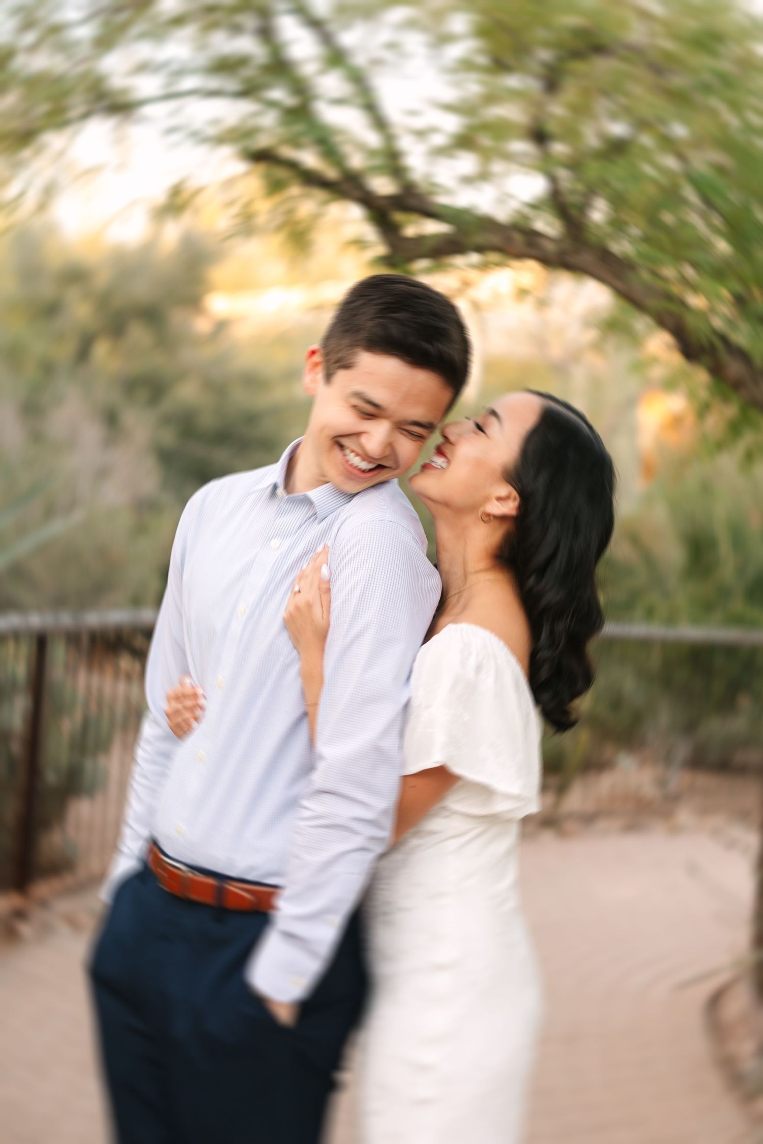 A romantic couple shares joyful laughter and embraces while taking engagement photos at sunset in a natural outdoor setting.