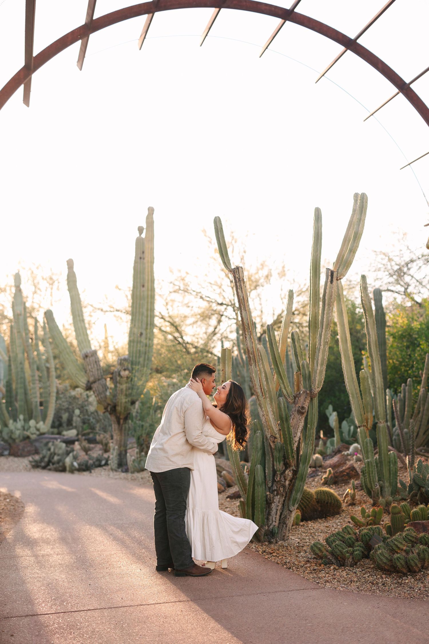 A romantic couple shares a tender embrace on a path surrounded by tall desert cactus at golden hour.