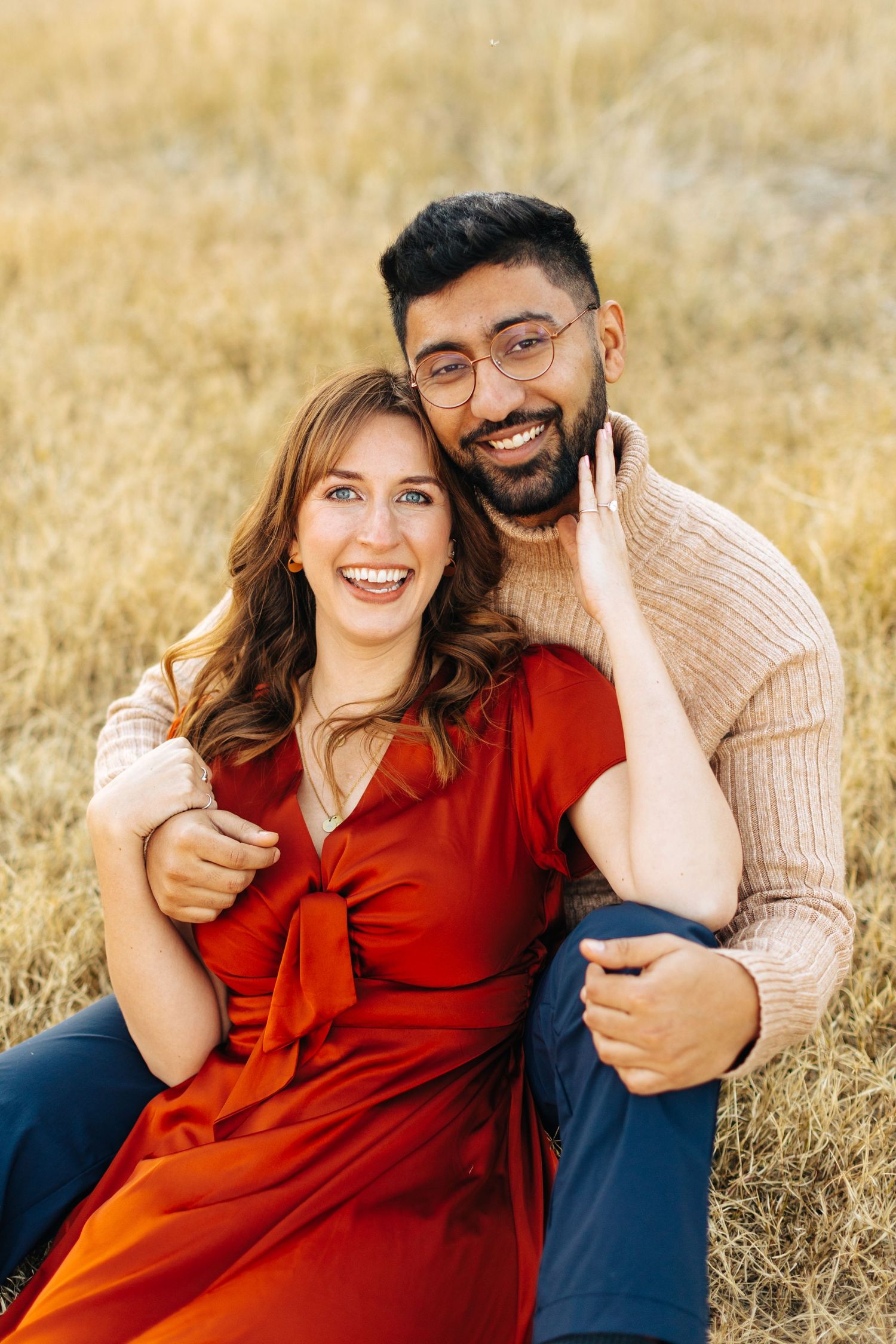 A couple sits together in a grassy field wearing autumn-toned clothing while sharing candid moments together.