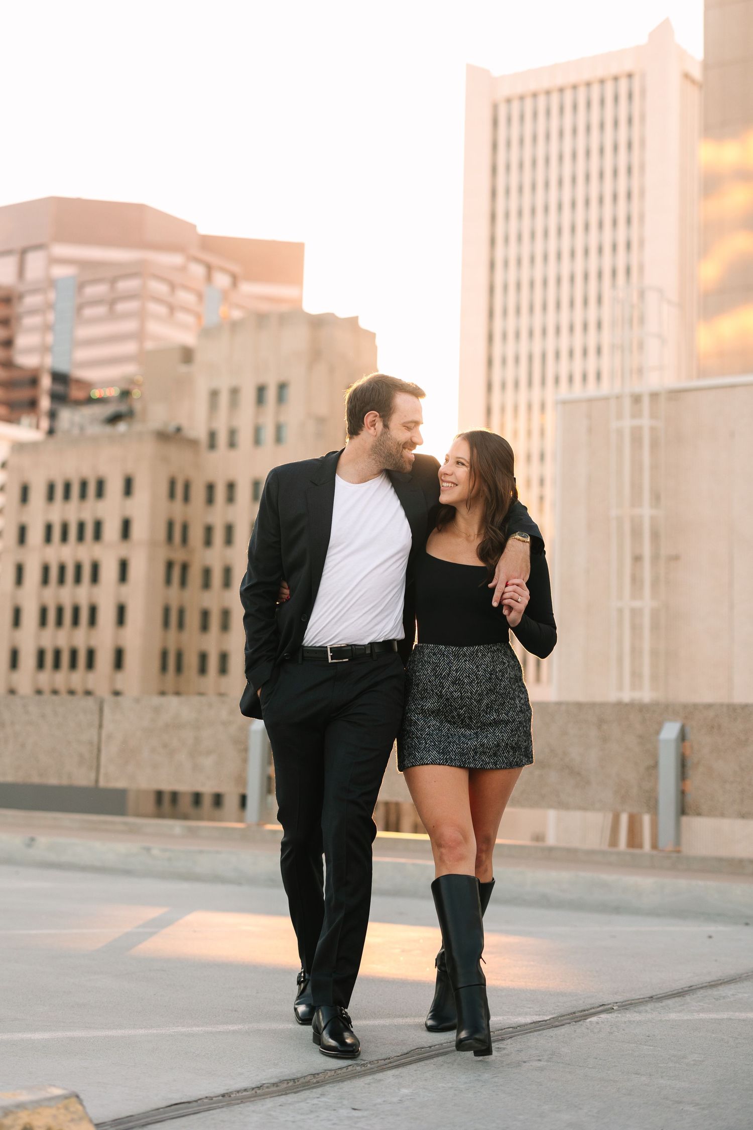 A couple dressed in black and white outfit walking together on a city rooftop during golden hour with skyscrapers behind them.
