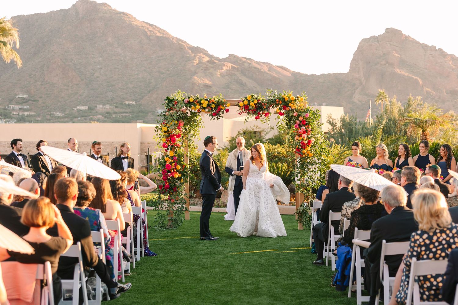 Outdoor wedding ceremony under a floral arch with mountain backdrop and rows of seated guests.