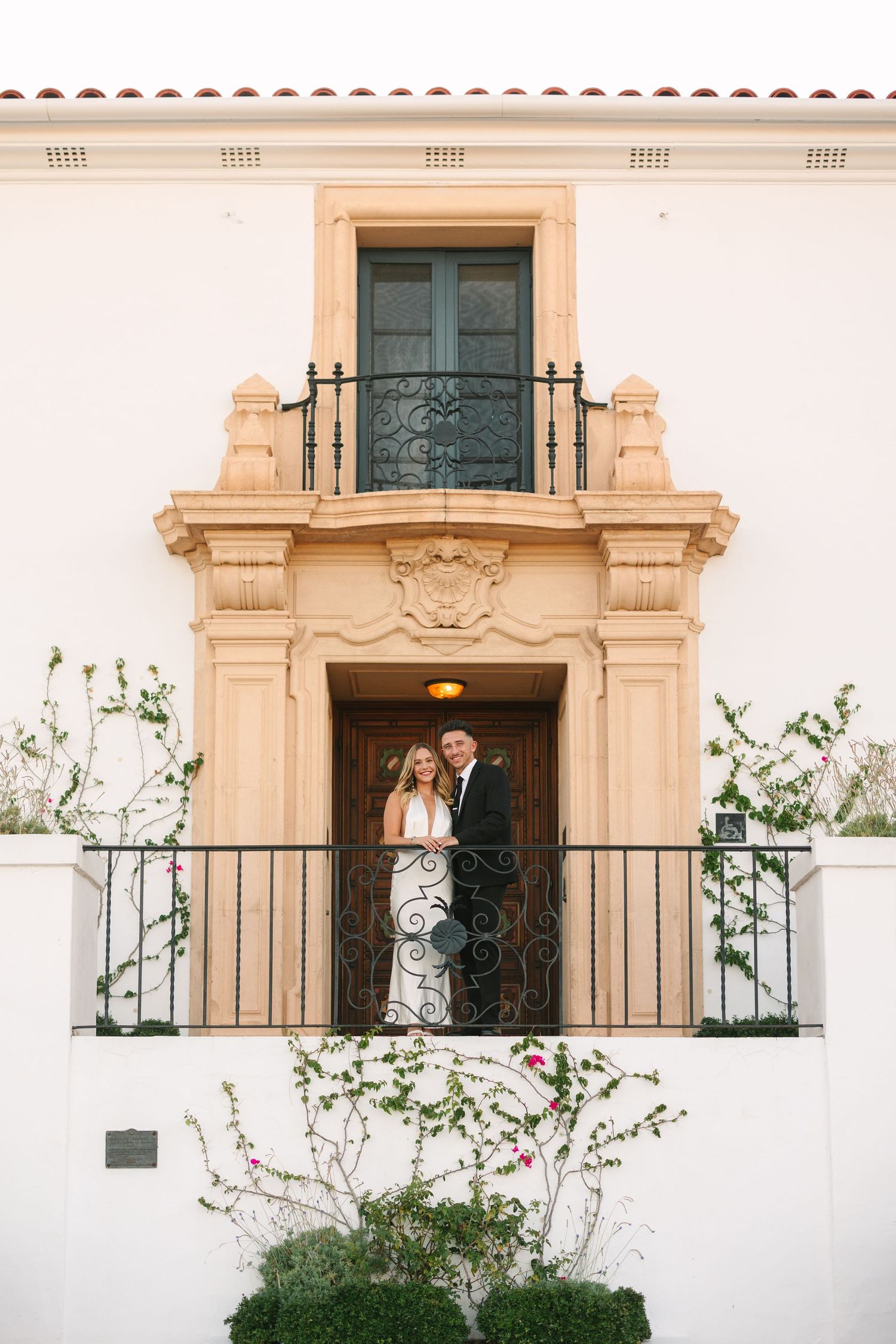 Two figures stand together on an elegant Spanish colonial balcony with ornate ironwork and cream-colored stone architecture.