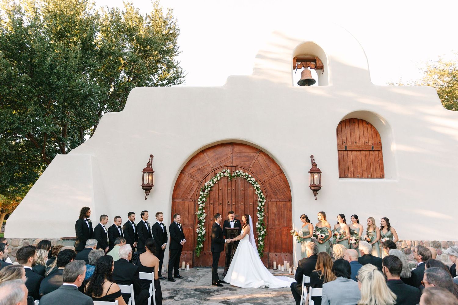 Wedding ceremony at white stucco chapel with arched wooden doors and guests seated.